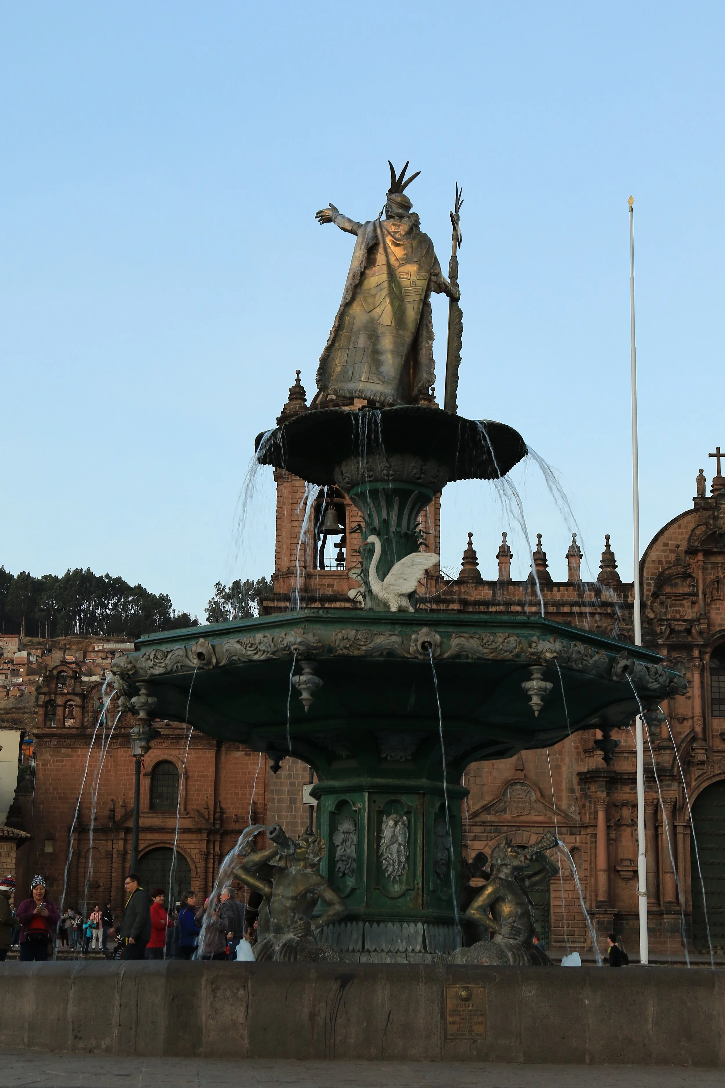 A large, ornate fountain with a statue of a man in traditional indigenous attire at the top, standing with one arm outstretched, in a public square with historic buildings and people nearby.
