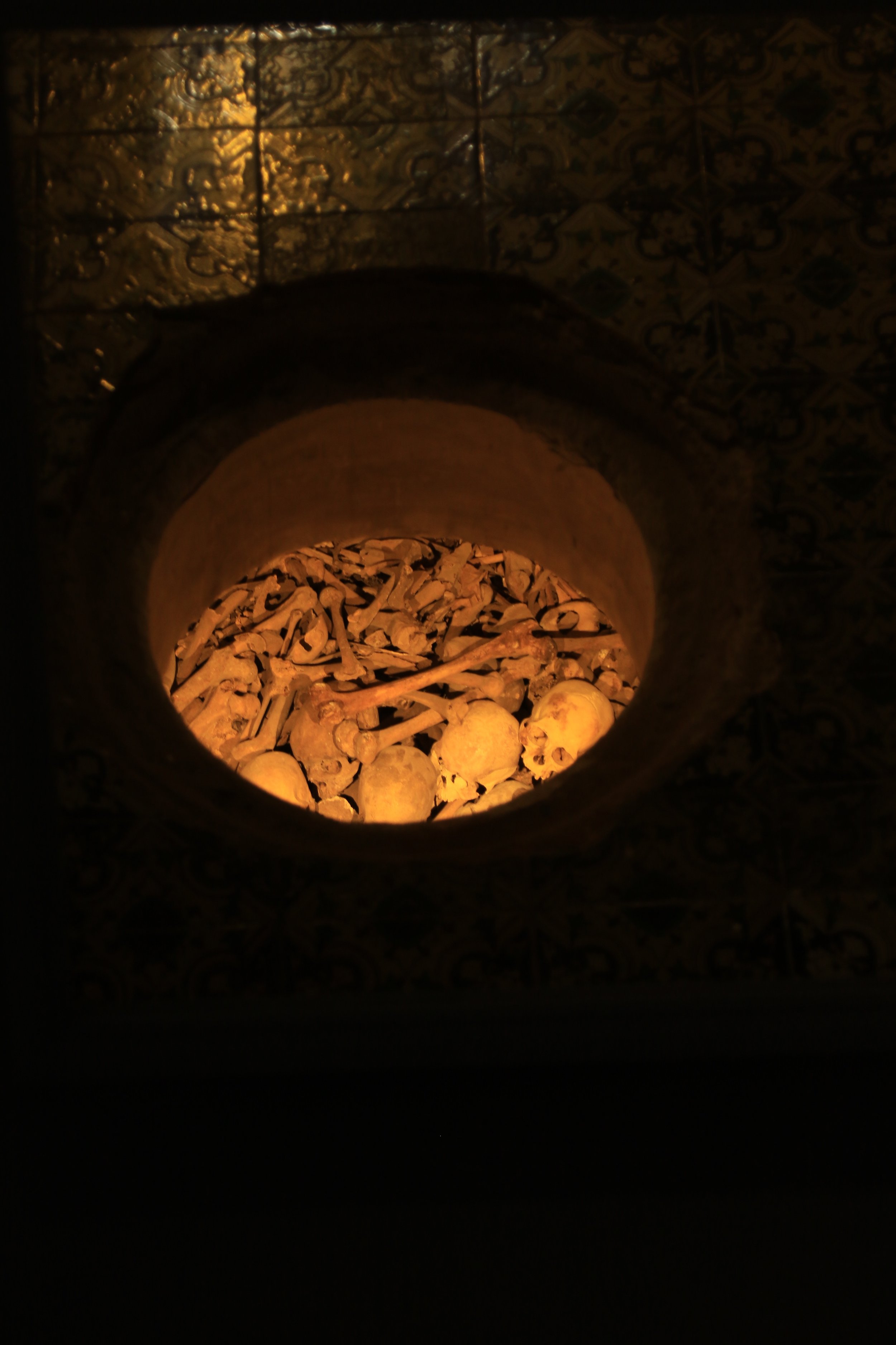 A view through a circular opening showing skulls and bones piled inside a dark, dimly lit space.