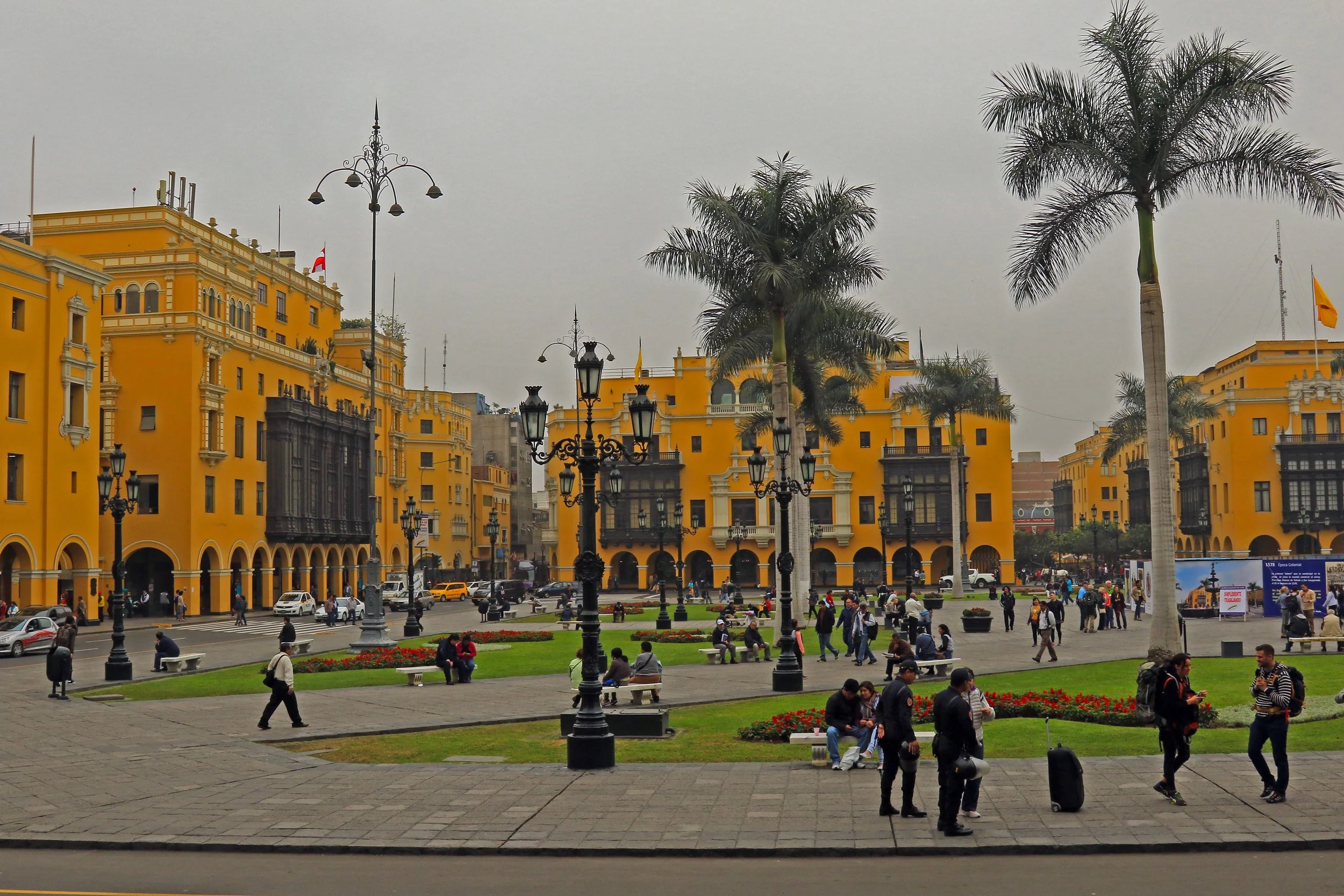 Urban city square with yellow colonial-style buildings, palm trees, benches, people walking and sitting, streetlamps, and a cloudy sky.