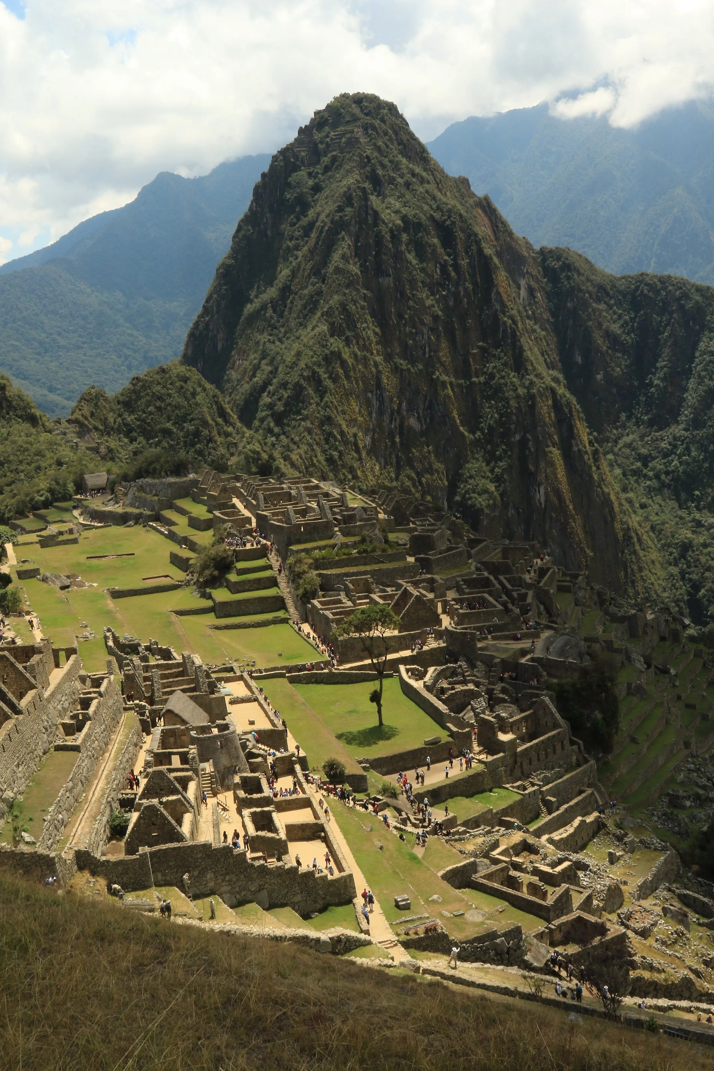 A panoramic view of Machu Picchu, an ancient Incan citadel in Peru, with stone ruins set among lush green terraces and a prominent mountain in the background under a cloudy sky.