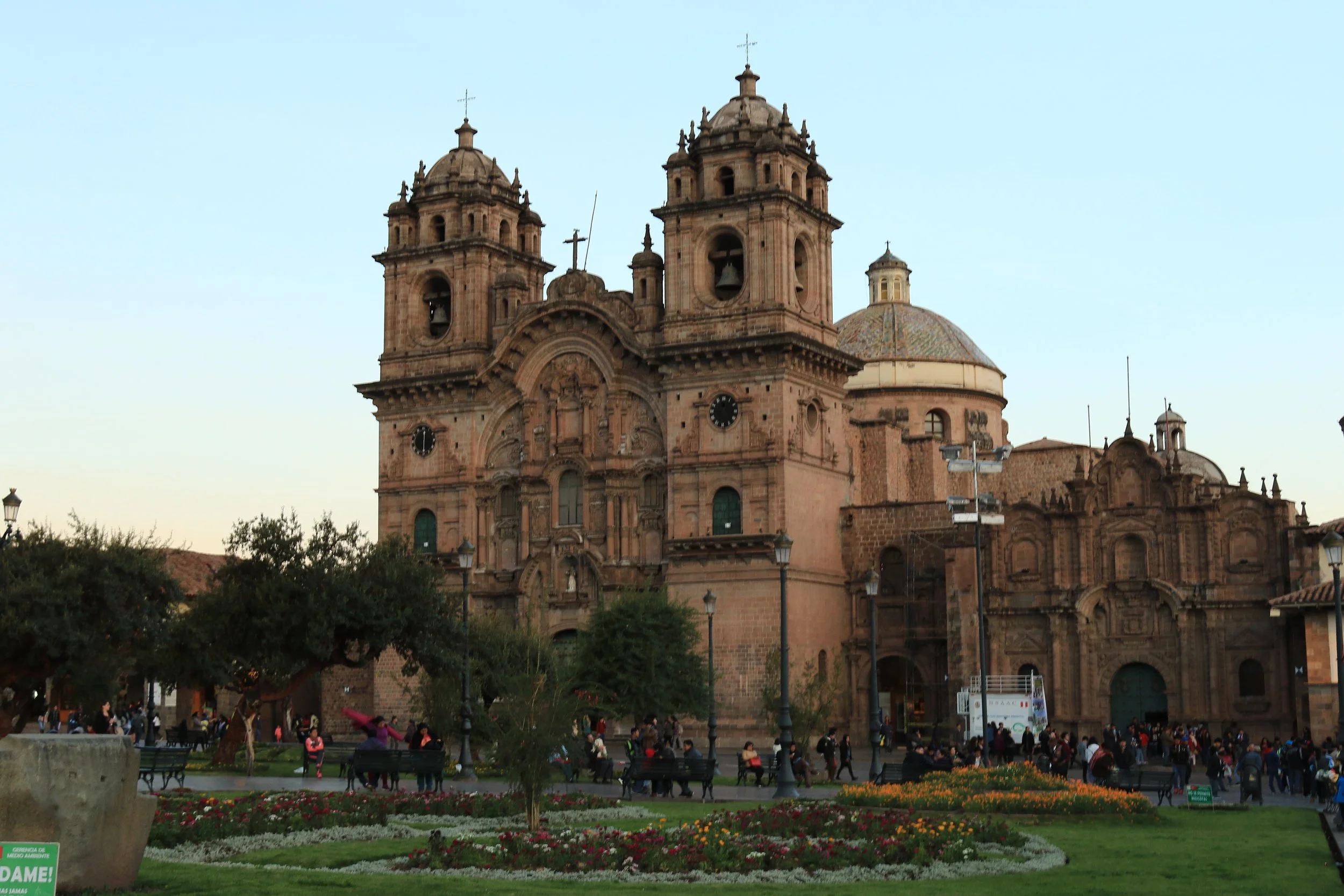 A historic church with twin bell towers, ornate architecture, and a rounded dome, situated in a busy square with people, park benches, trees, and flowers