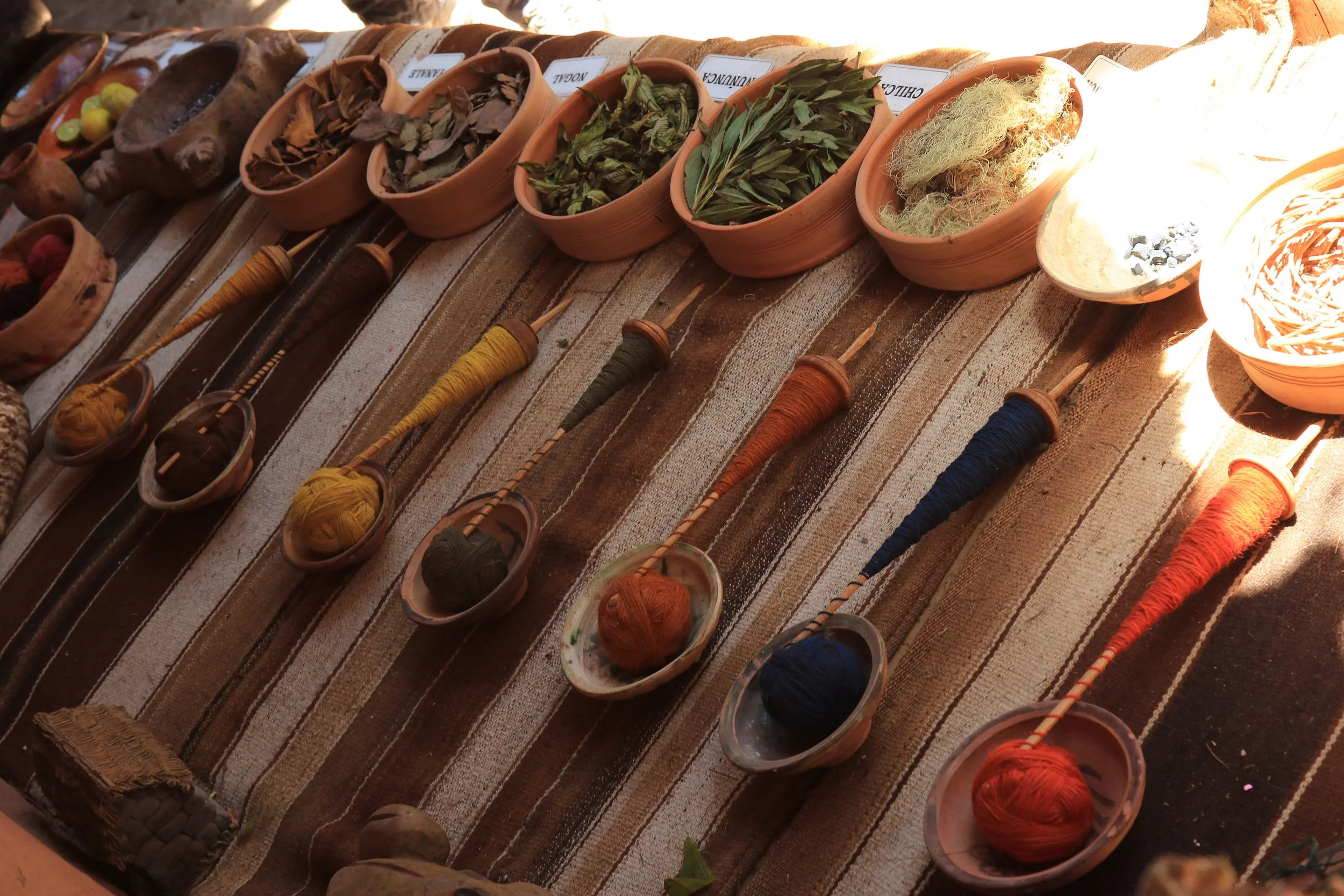 Colorful spools of thread arranged on a striped fabric, with bowls of leaves and other natural materials behind them, in a marketplace setting.