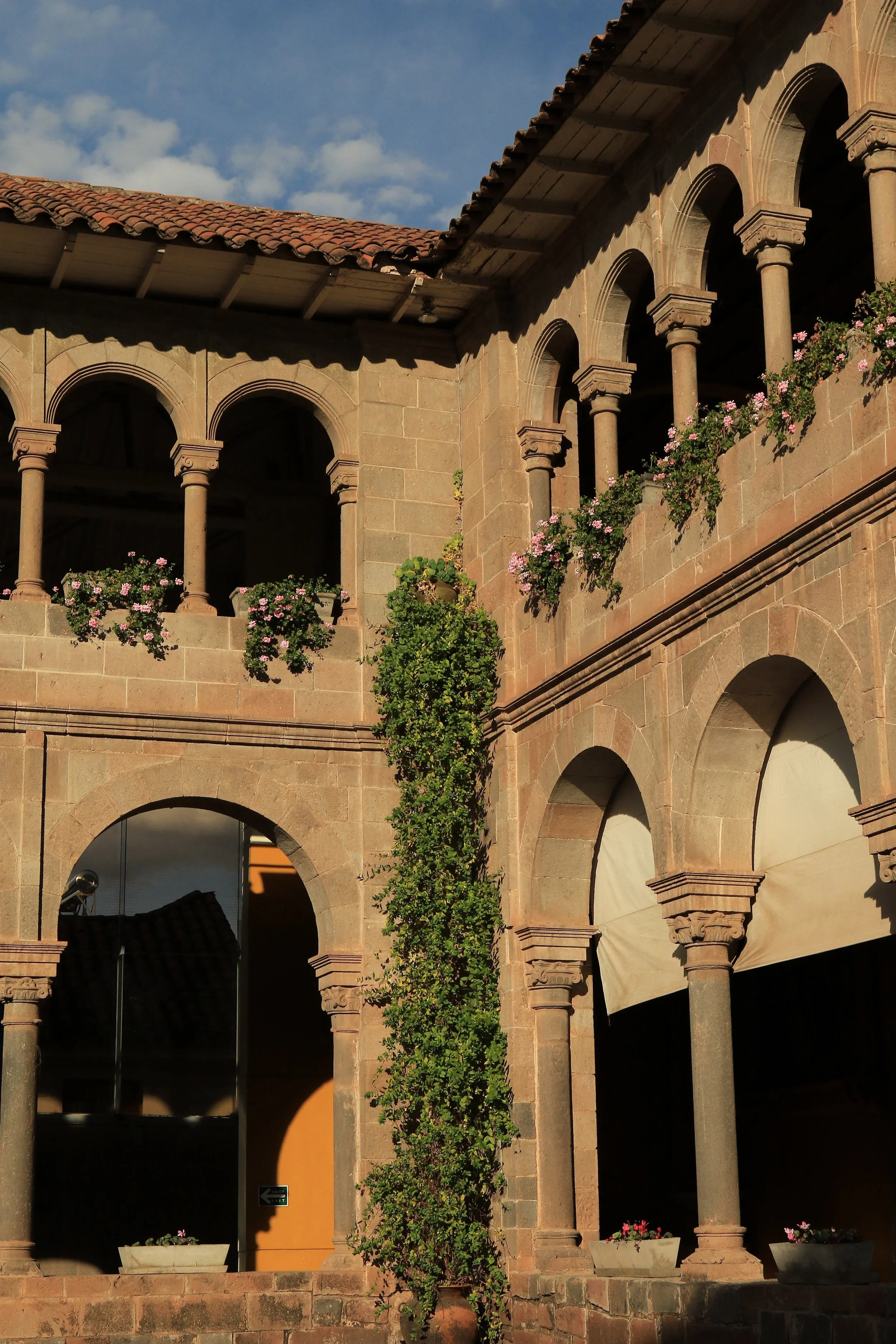 Historic stone building with arched windows and columns, decorated with pink and white flowers on the upper balcony, under a blue sky with clouds.