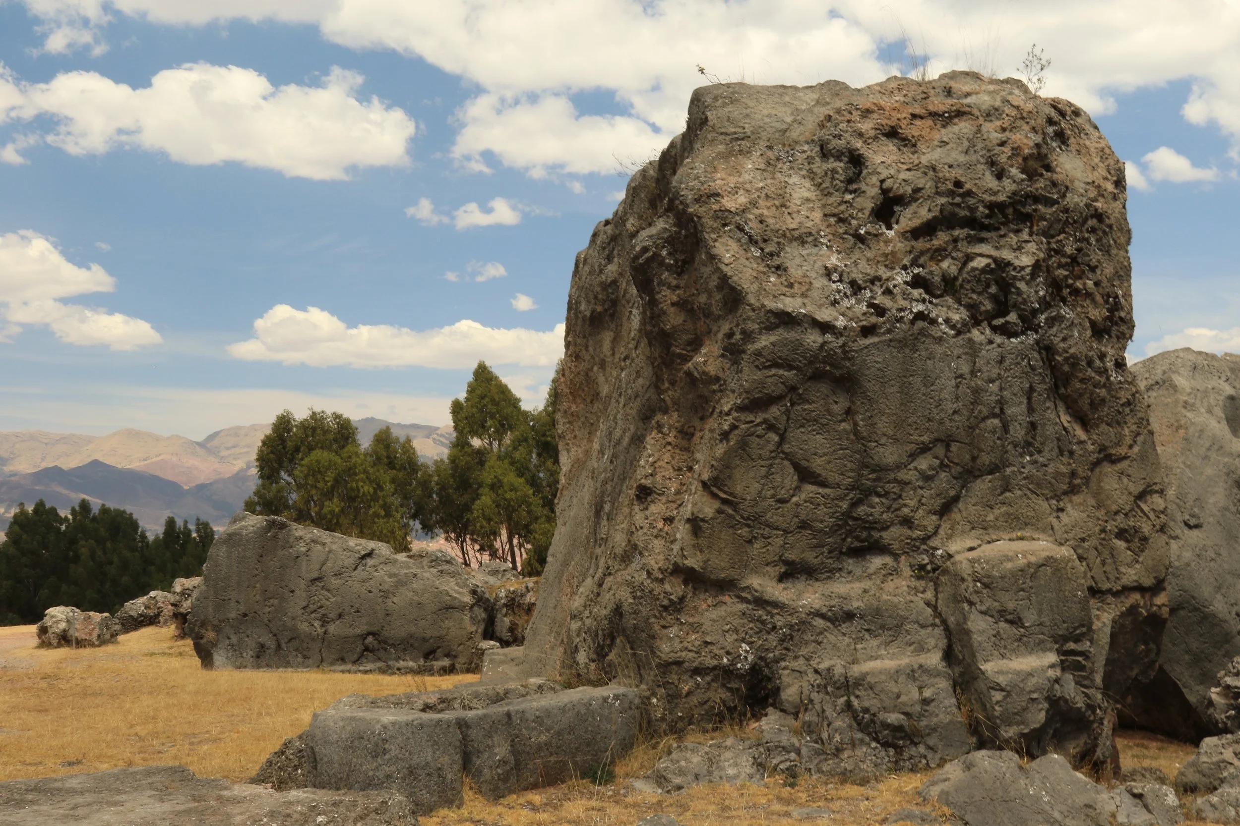 Large rocks and boulders in a field with trees, mountains, and a blue sky with clouds in the background.