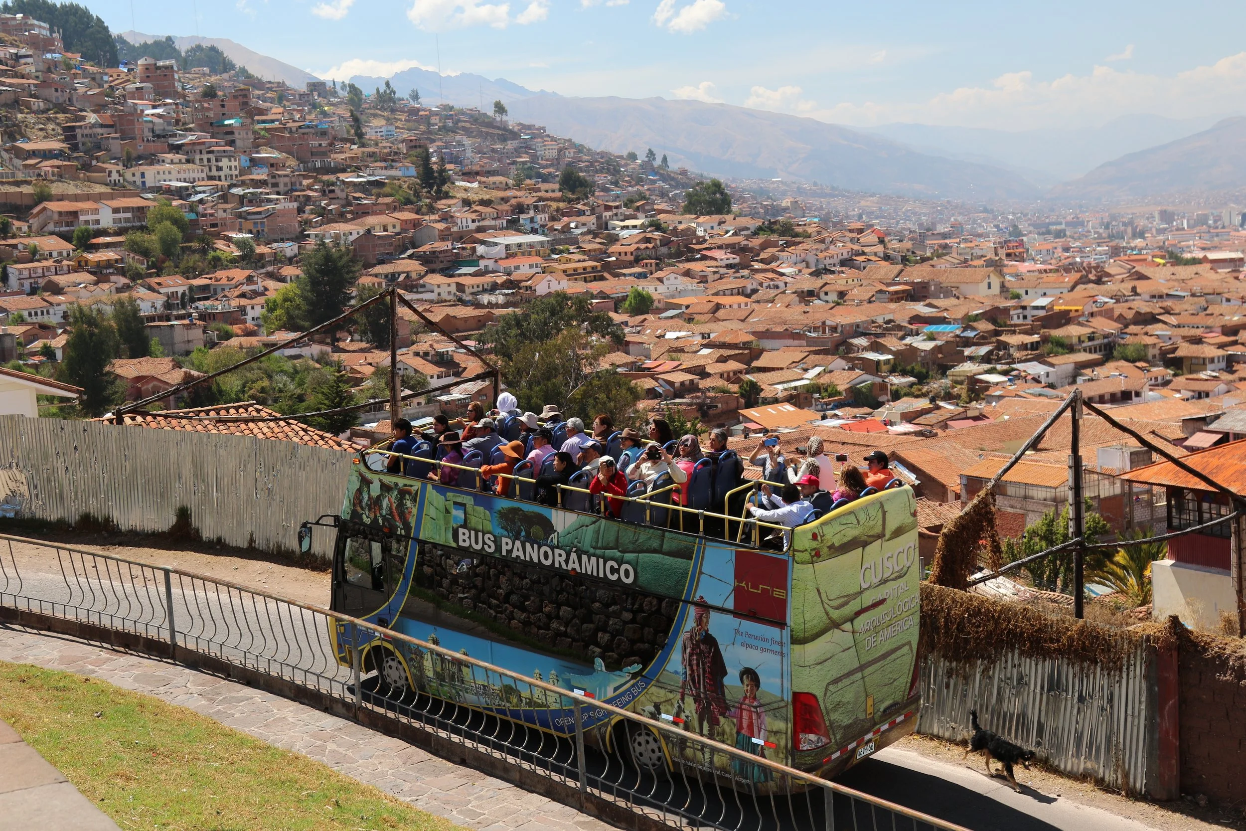 An open-top double-decker bus filled with tourists riding through a hilly urban area with many houses and mountains in the background.