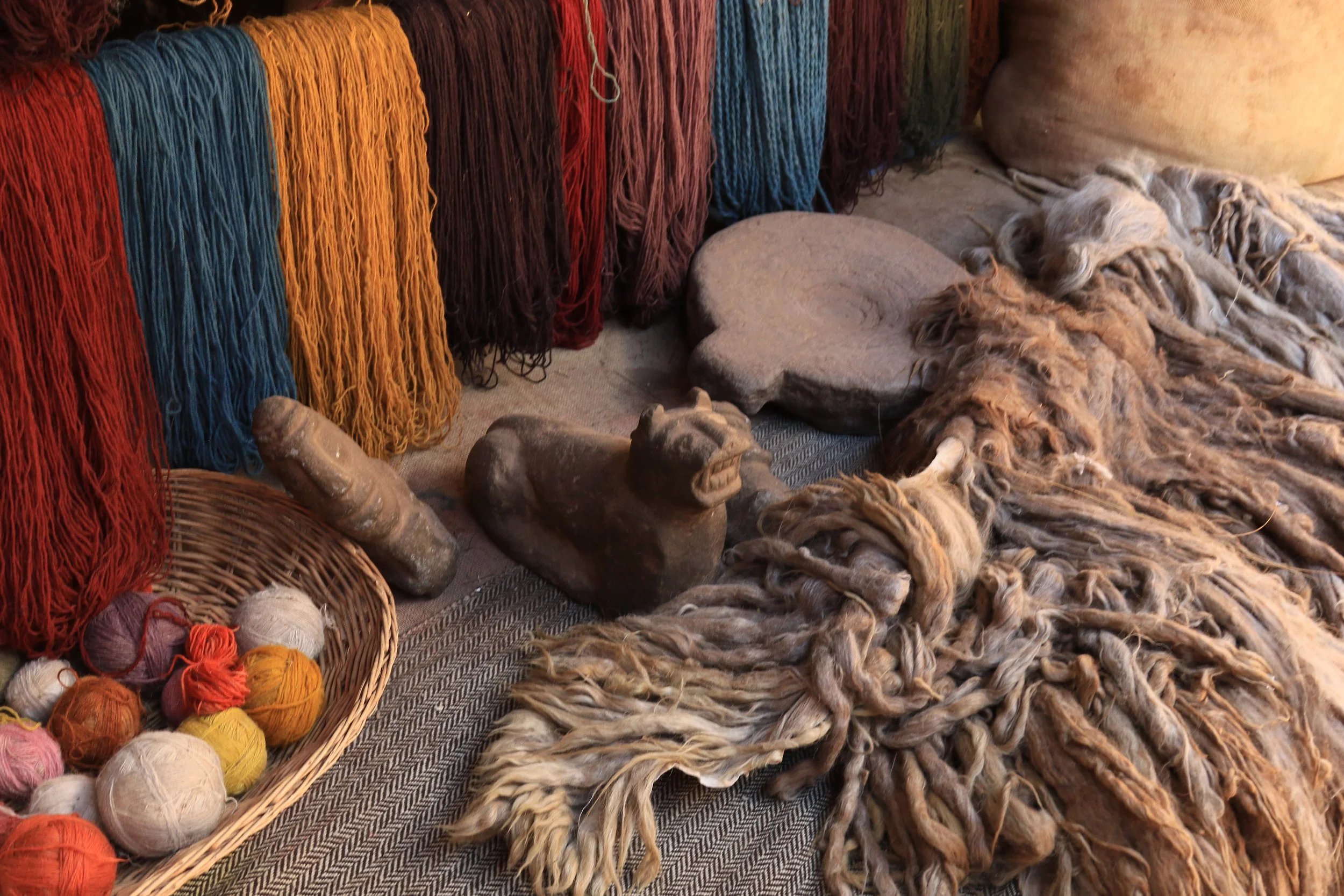 Colorful balls of yarn, woven basket, carved stone sculptures, and woolen textiles with long, twisted fibers on a fabric surface.