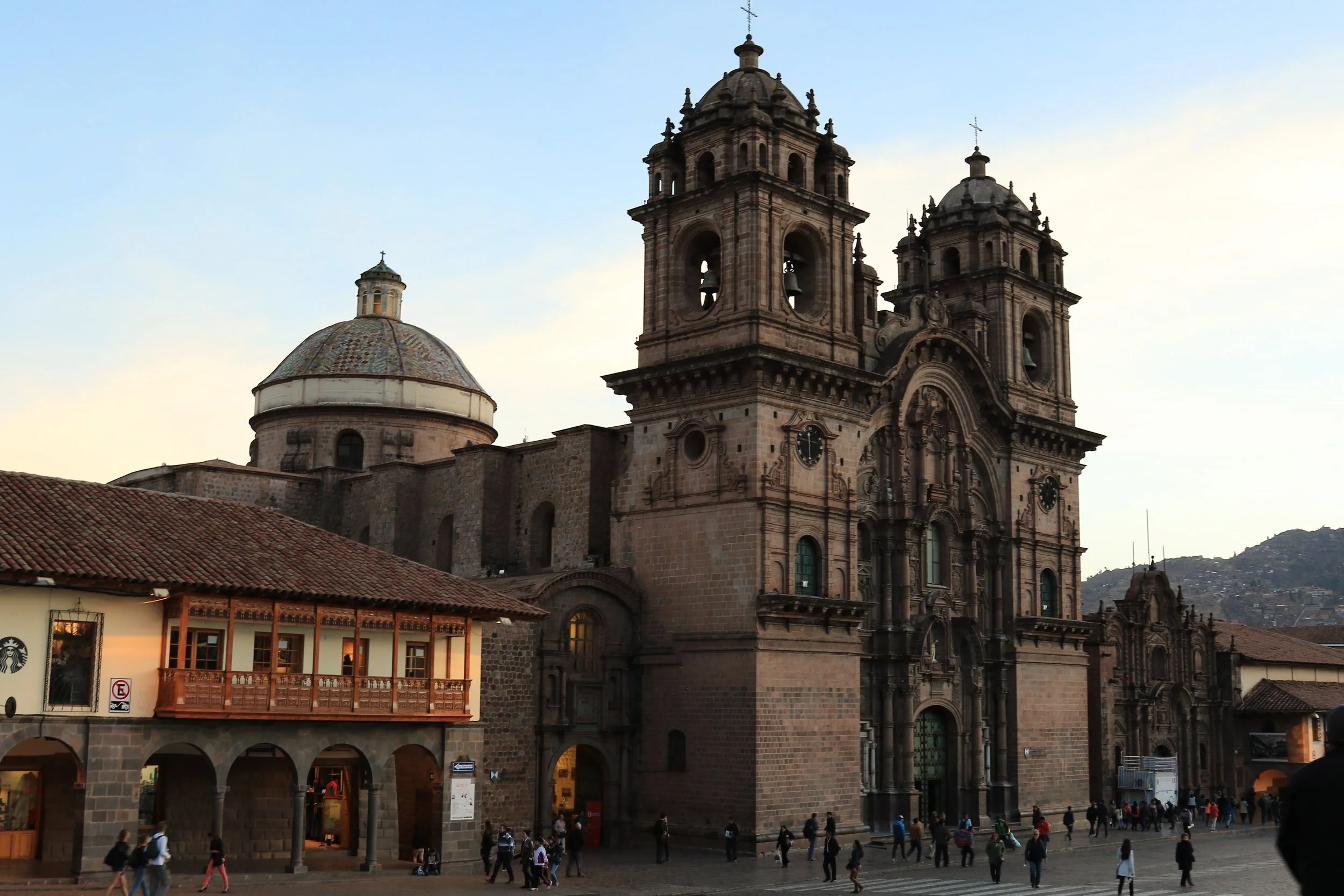 Historic cathedral with two tall bell towers and a large dome, situated in a busy plaza with people walking and a small building with a balcony in the foreground.