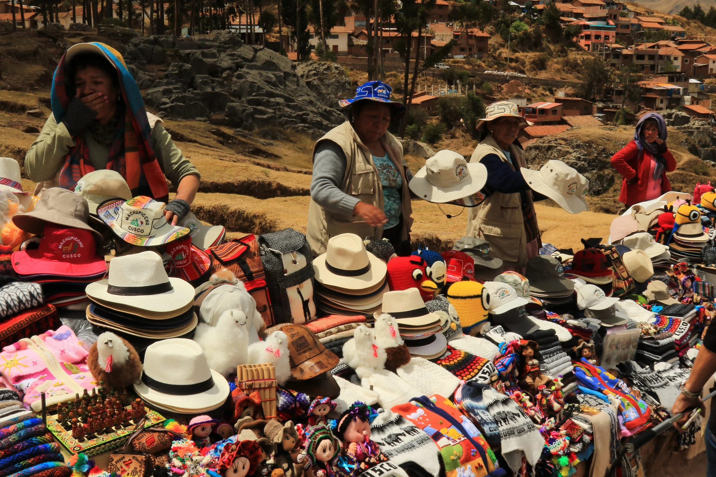 Market stall displaying hats, plush toys, knitted items, and souvenirs with women selling merchandise outdoors on a hillside, houses visible in the background.
