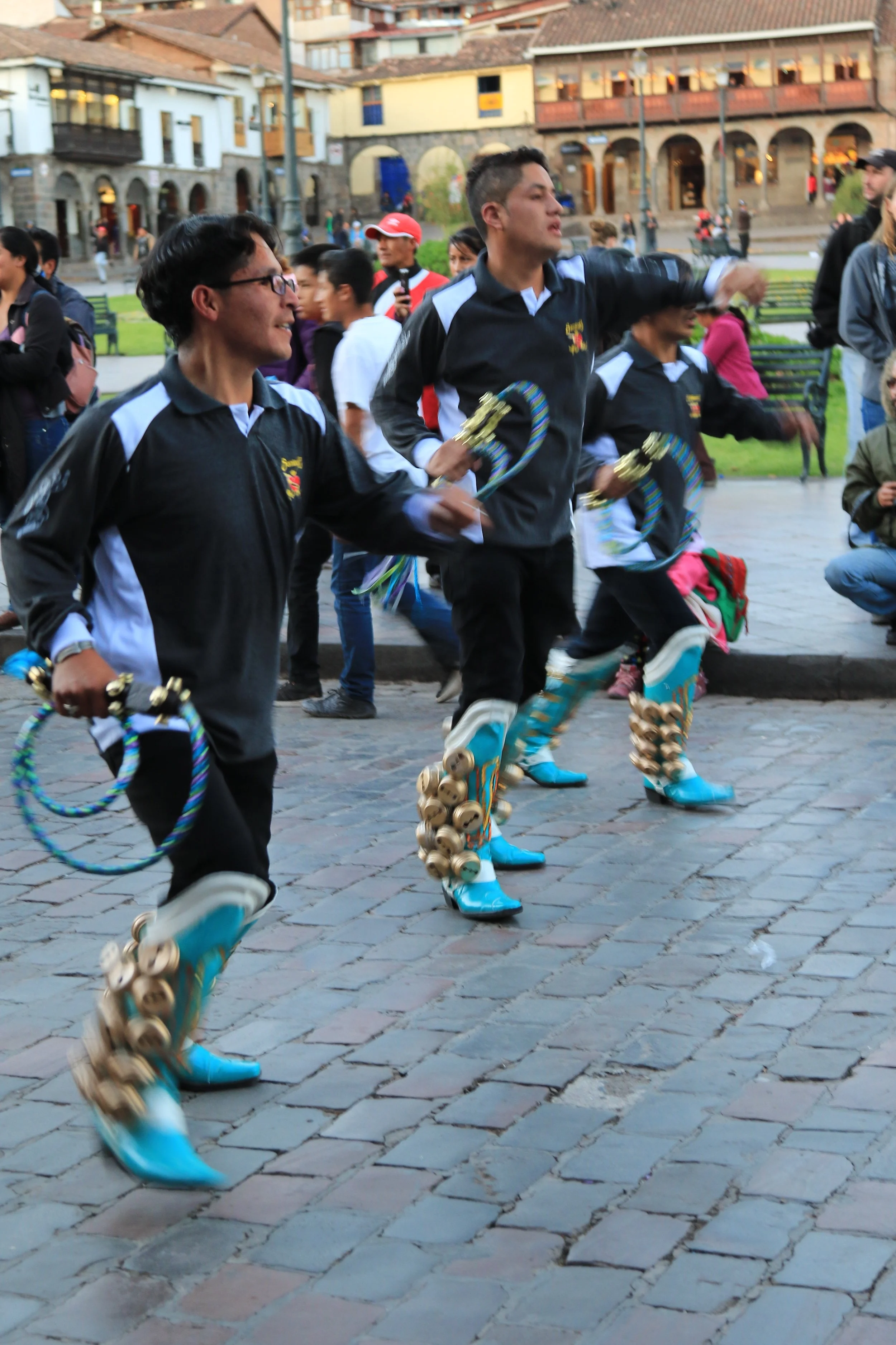 Three performers in traditional attire dancing with bells on their boots and holding colorful hoop-like props in a public square with onlookers and old buildings in the background.