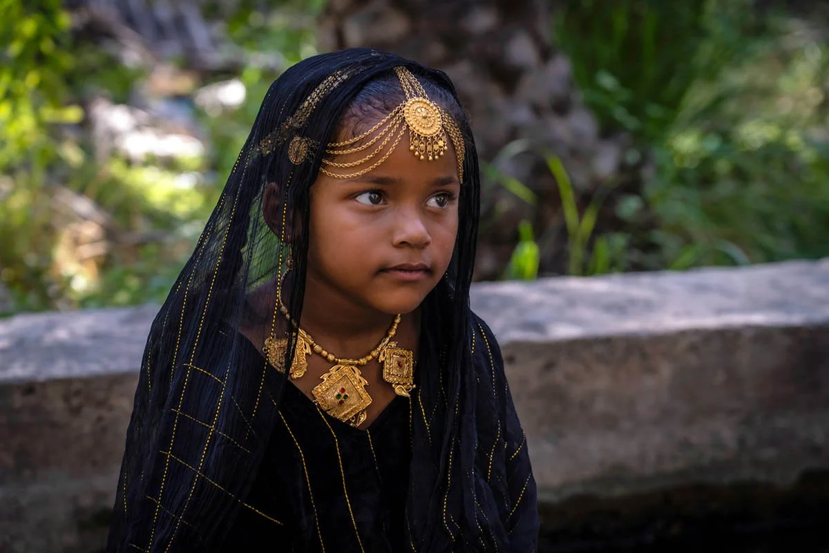 Young girl wearing traditional jewelry and a black netted veil, outdoors with greenery background.