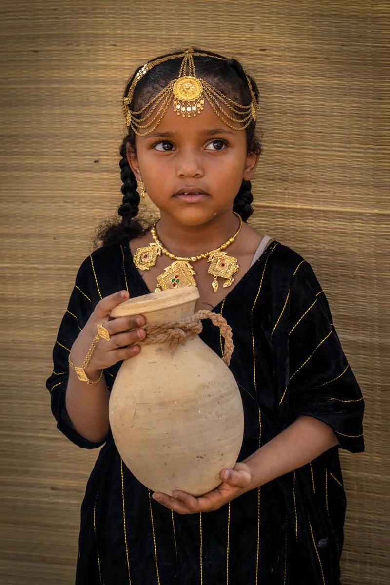 A young girl dressed in traditional clothing with gold jewelry, holding a clay vessel with a lid, standing against a woven background.
