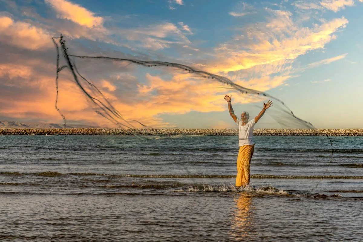 A person standing in the ocean with arms raised, casting a fishing net during sunset with colorful clouds in the sky.