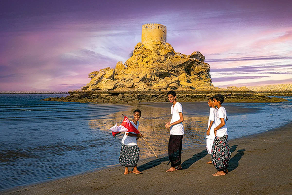Four children standing on a beach, with a rocky island or fortress in the background and a pink and purple sky. Two children are smiling, one is holding a flag, and they are all dressed in traditional clothing.