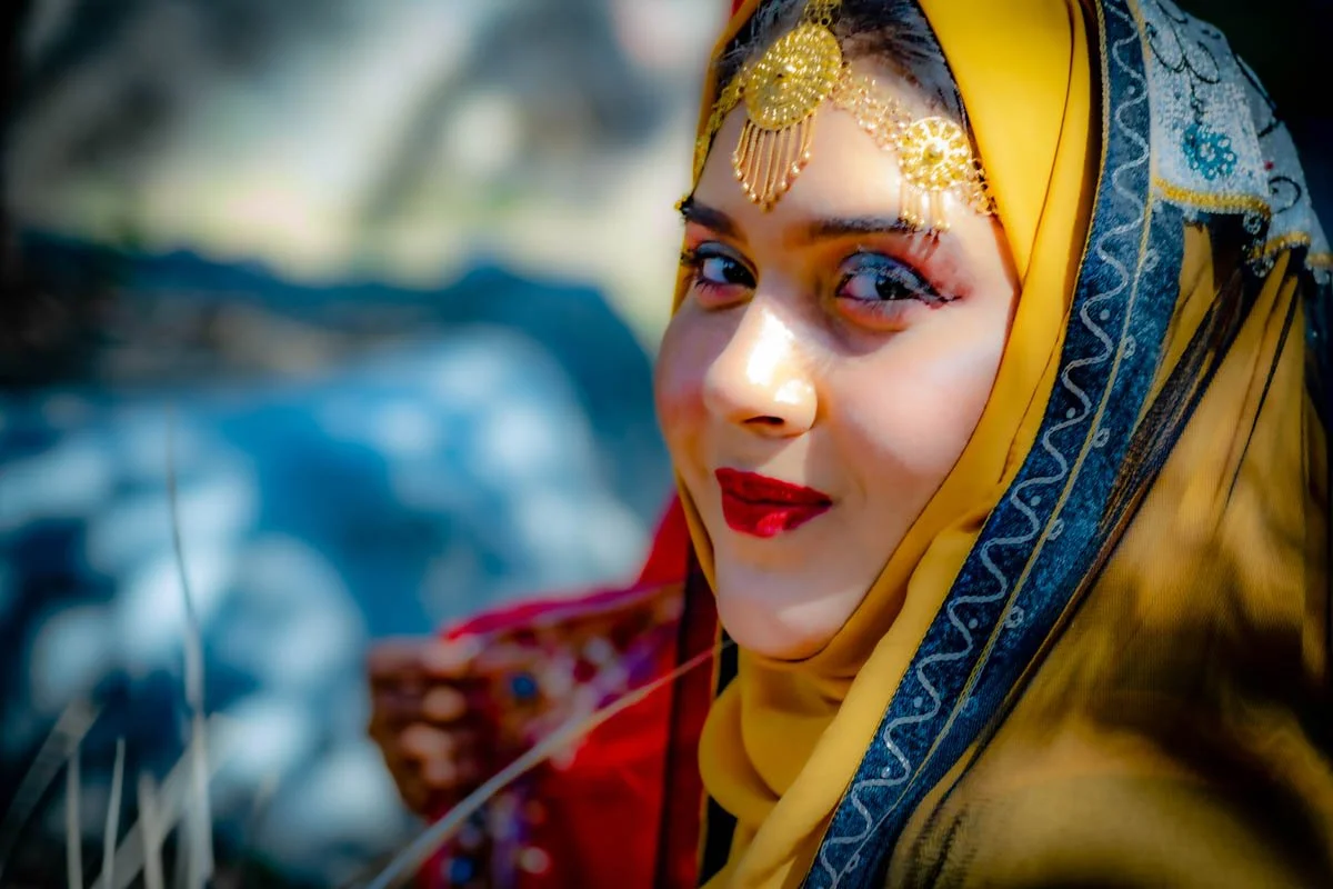 A woman wearing a yellow headscarf with blue and gold embroidery, gold jewelry on her forehead, and bright red lipstick, smiling at the camera.