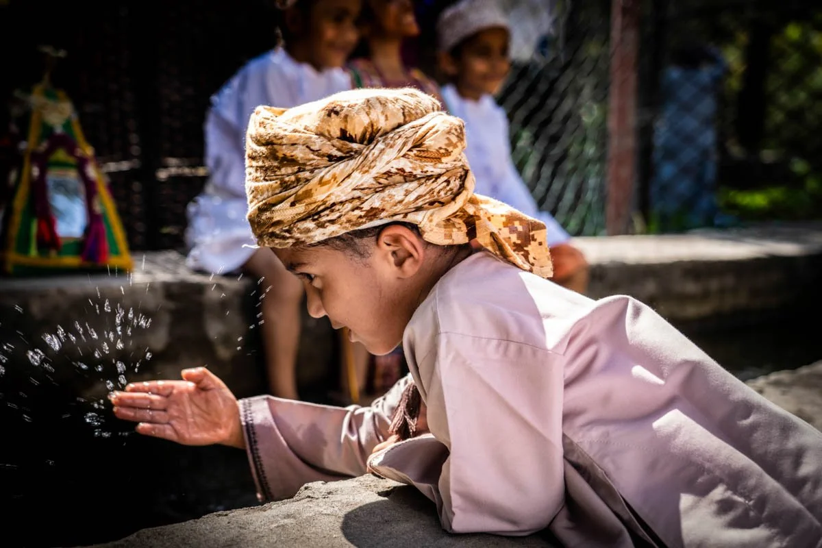 A young boy in traditional attire with a headscarf leans over a stone surface, splashing water from his hand, with three children sitting and smiling in the background.