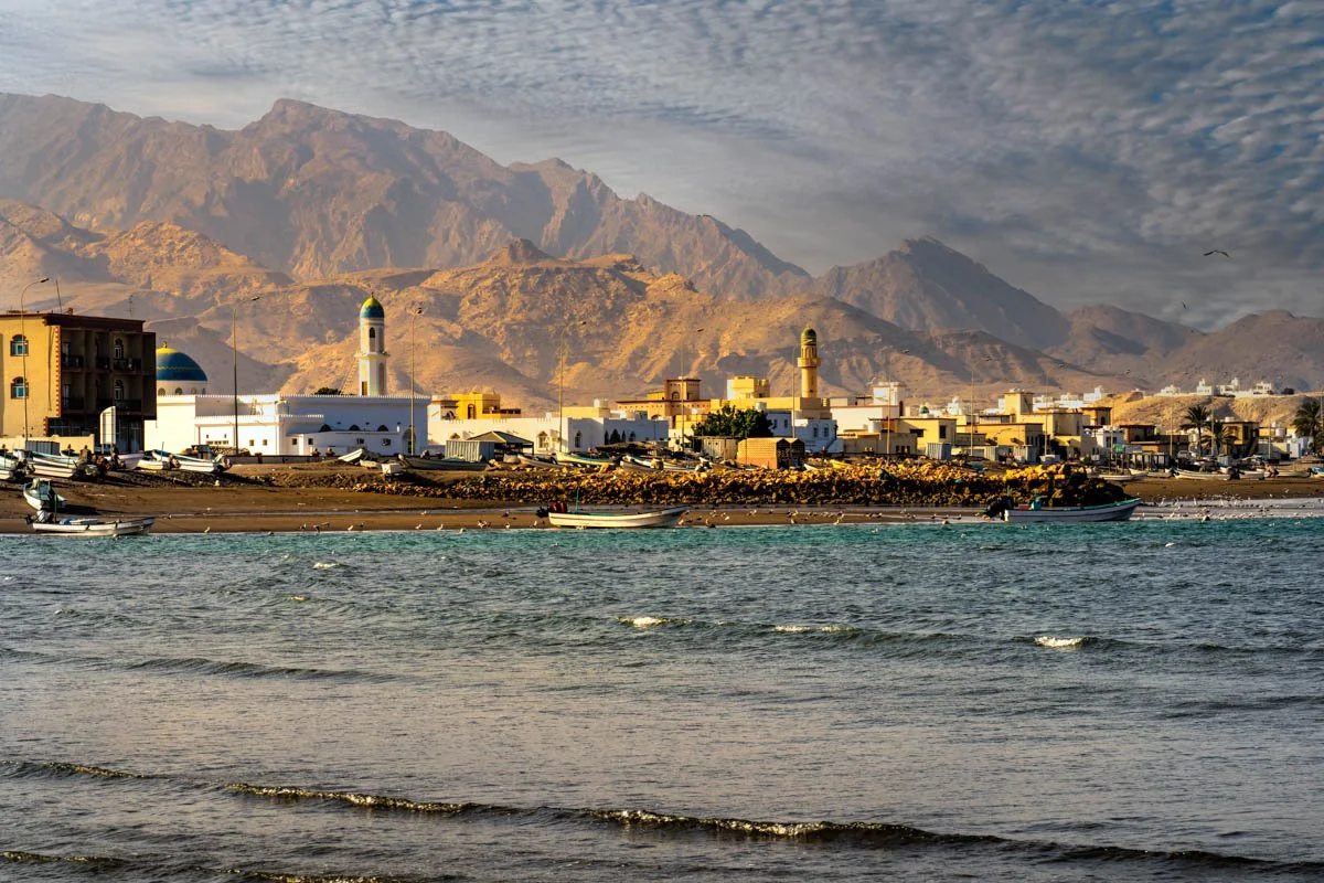 A coastal town with white buildings and mosques with domed roofs, set against arid mountains in the background. The waters in the foreground have small boats docked along the shore, and a partly cloudy sky is visible above.