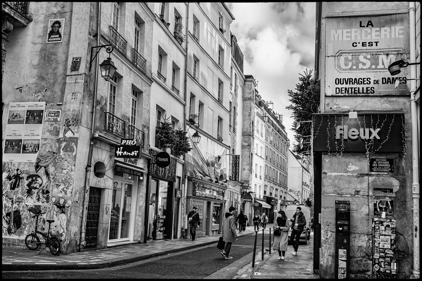 A street scene in Paris with pedestrians walking along the sidewalk, lined with shops and apartment buildings. Signs for Vietnamese cuisine and various stores are visible, with some posters and graffiti on the walls. The sky is partly cloudy.