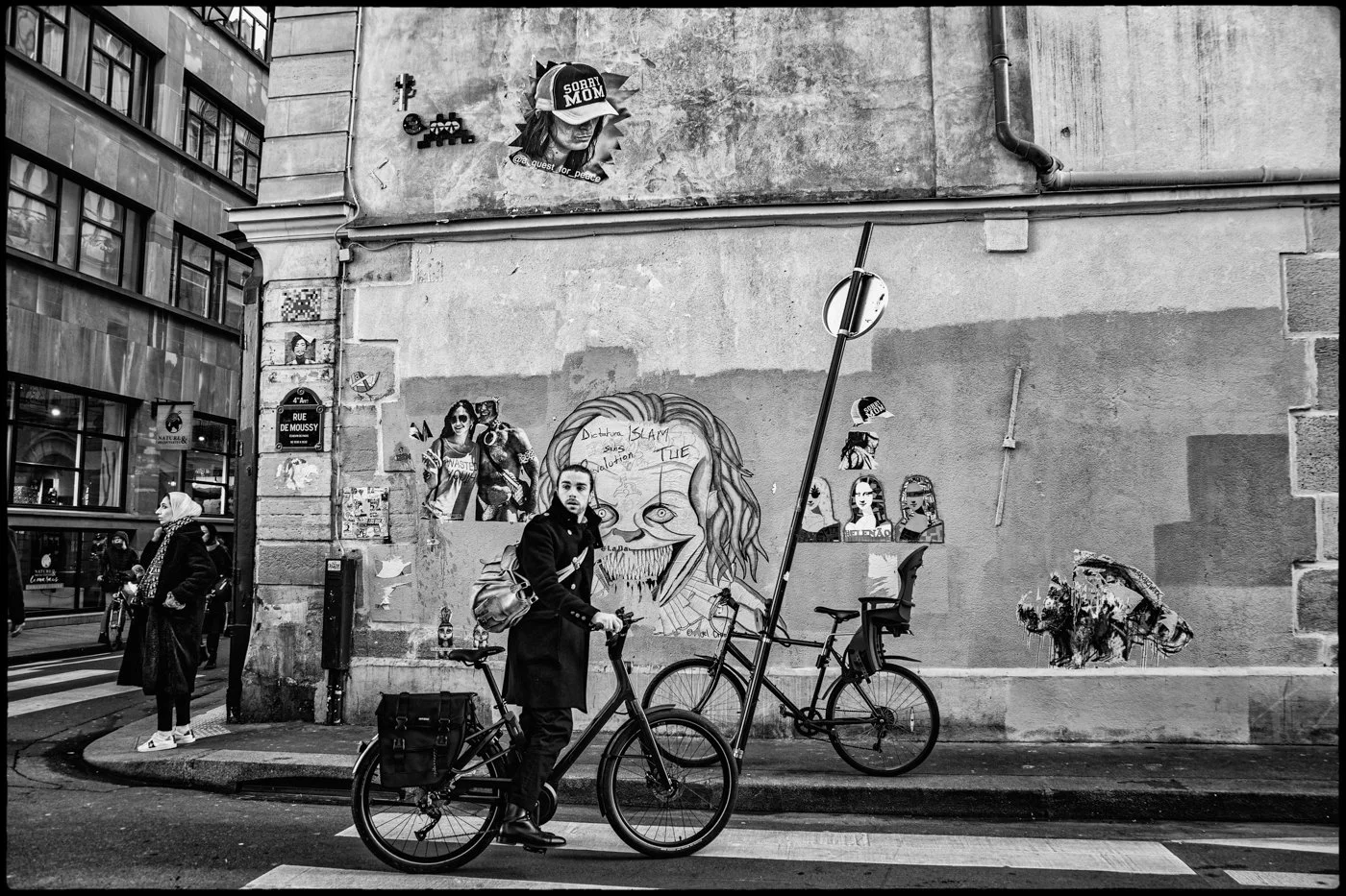 A man with a backpack stands next to a bicycle at a street corner in an urban setting, with street art and posters on the wall behind him, including a large graffiti face with the words "Dictadura BLAM" and "TUE" written on it.