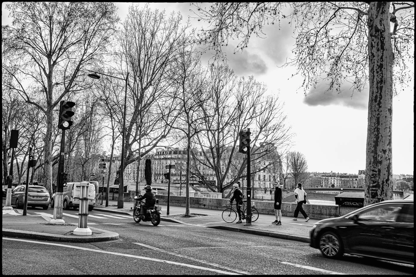 City street scene in black and white with traffic lights, cars, pedestrians, and leafless trees by a river with buildings in the background.