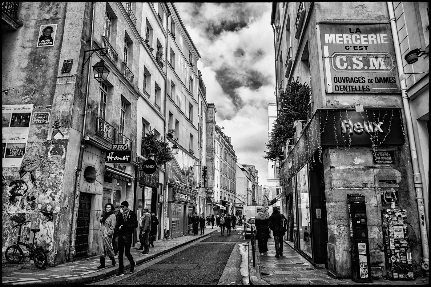 Street scene in Paris, France with people walking along a narrow street lined with shops and cafes, old buildings, and cloudy sky.