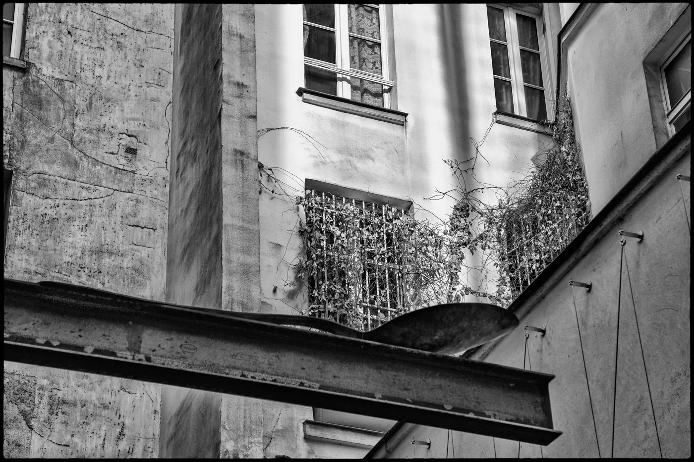 Black and white photograph of an old building's interior courtyard with three windows, some of which are covered with ivy, and an angled metal beam in the foreground.