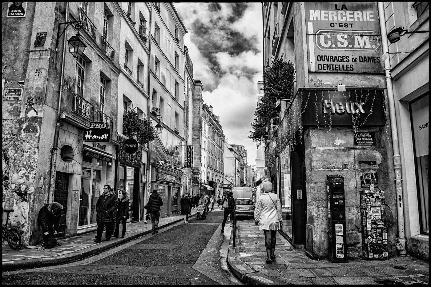 Black and white photo of a city street with people walking. There are storefronts and signs, including 'PHO', 'Hanoi', 'Floux', and a vending machine. Buildings are tall with balconies, and the sky is cloudy.