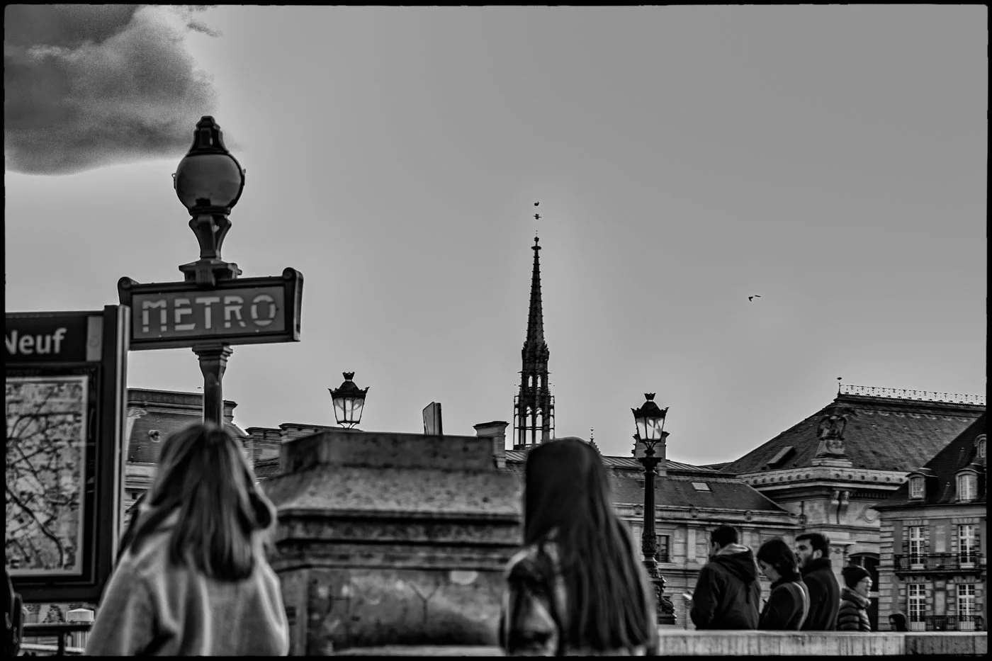 People waiting near a metro entrance on a Paris street, with historic buildings and a church steeple in the background, under a cloudy sky.