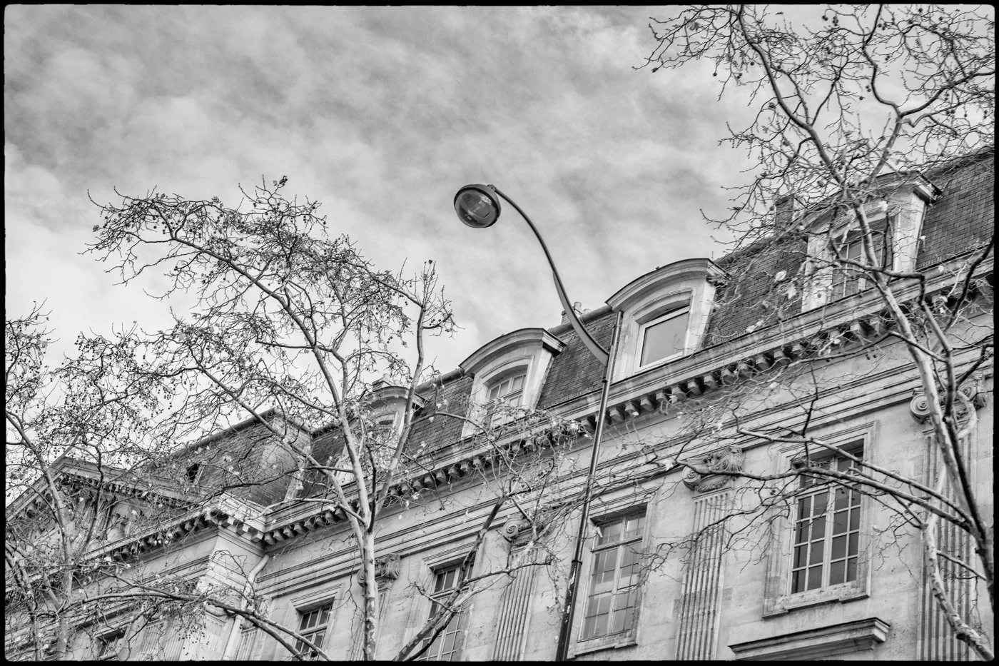 Black and white photo of an old building with large windows, decorative architectural details, dormer windows, and a streetlamp, with leafless trees in the foreground, against a cloudy sky.