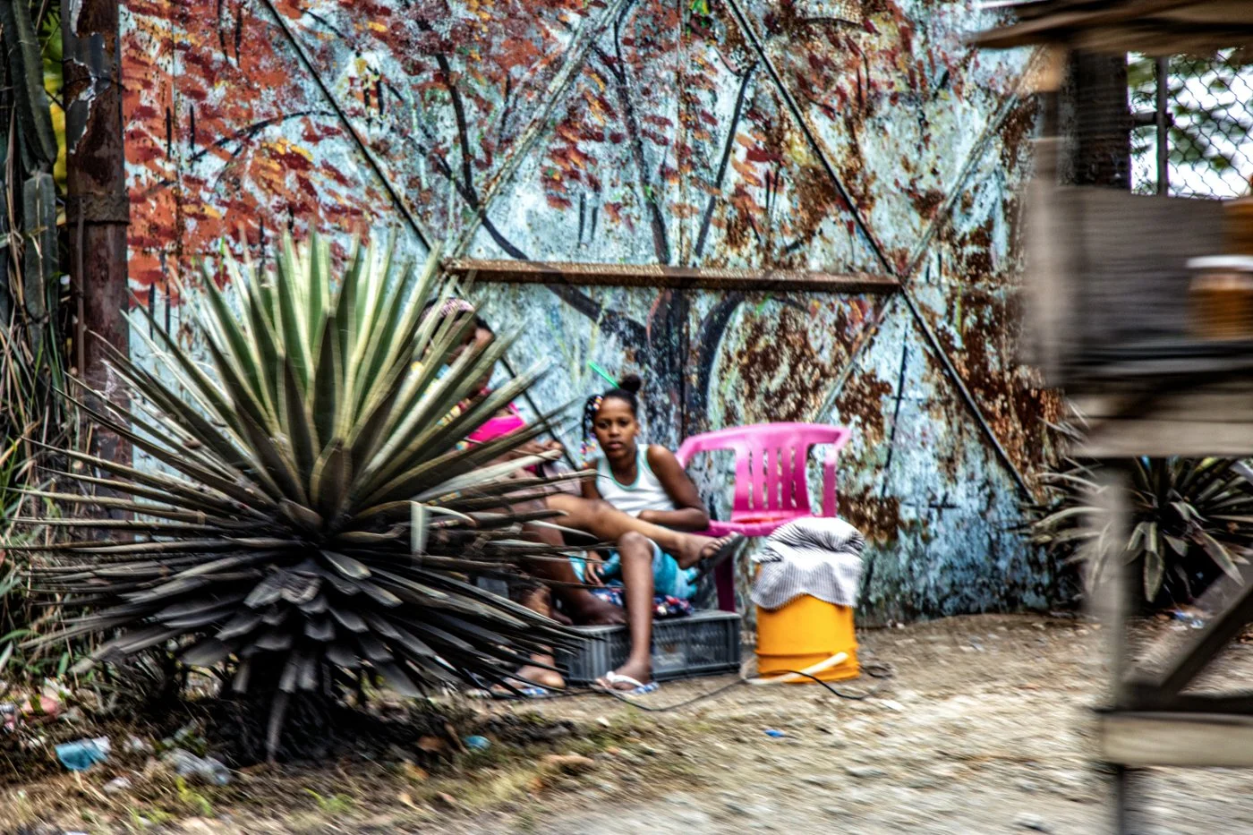 Young girl sitting on a pink plastic chair placed on a yellow container, near a large spiky plant and a colorful wall with graffiti, in an outdoor setting with dirt ground.