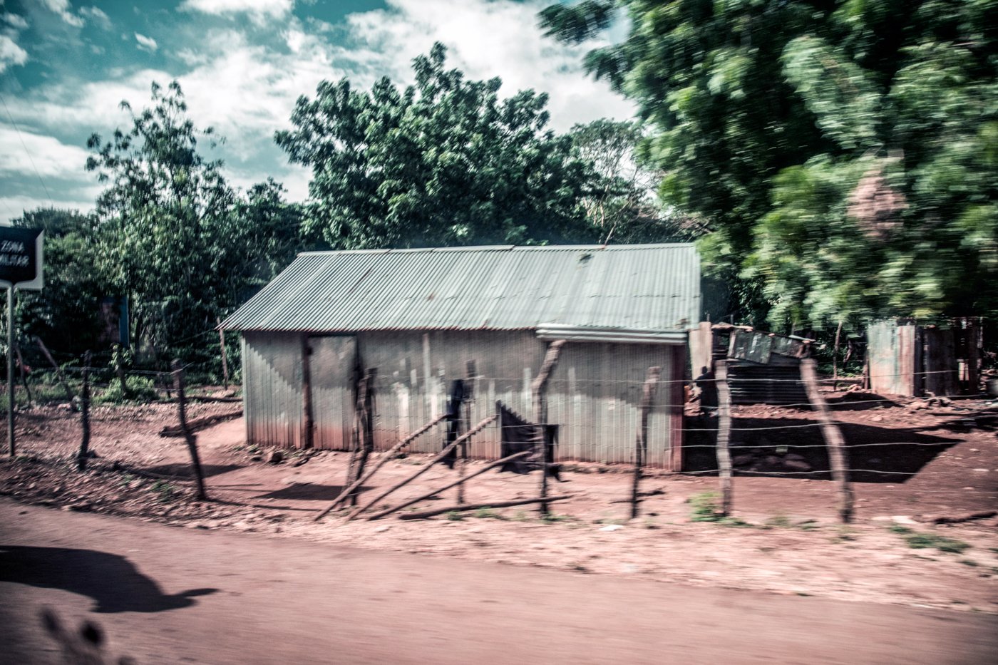A small, rustic house with a metal roof, surrounded by a wooden fence and trees, on a dirt road.