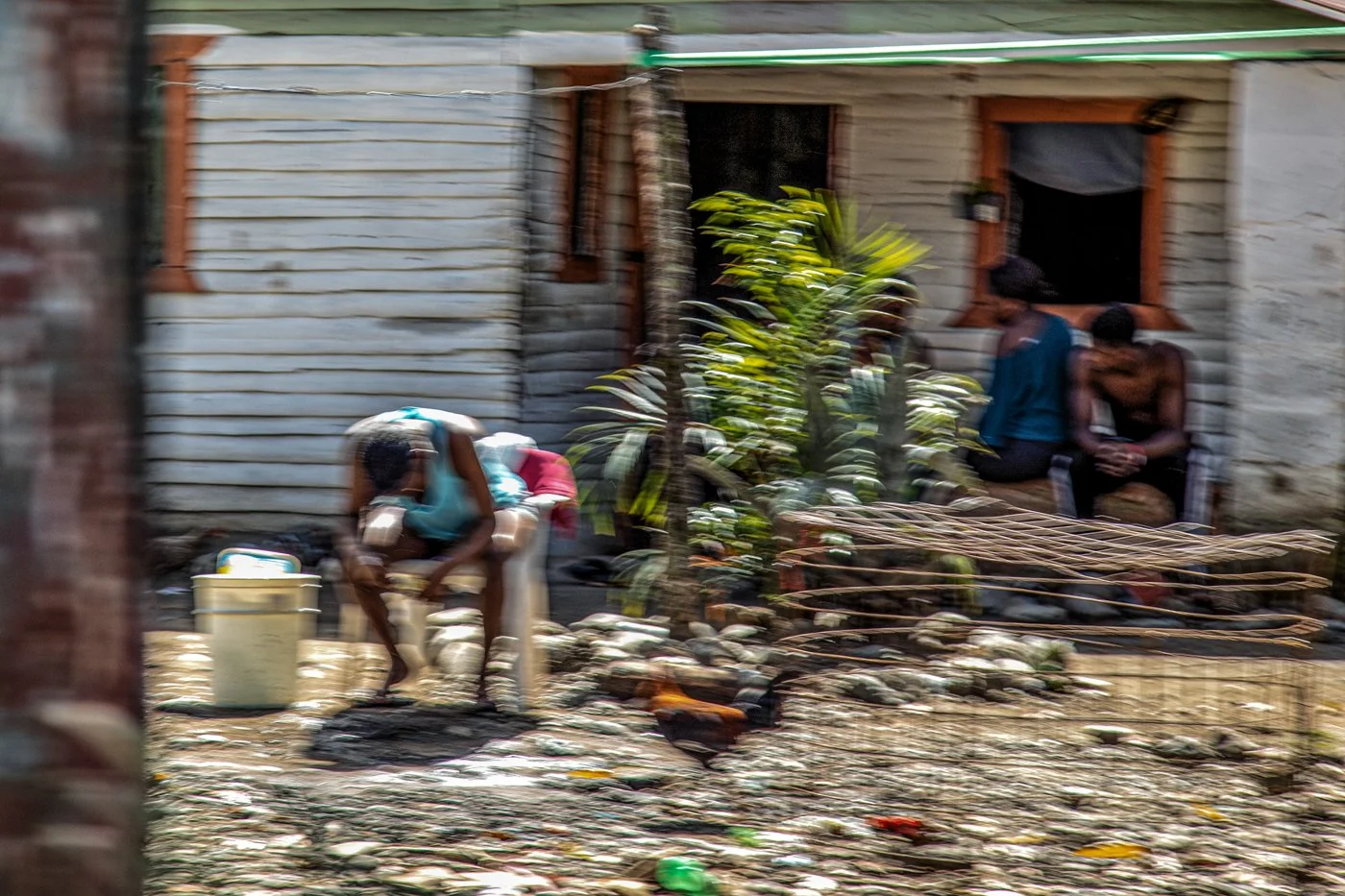People sitting and standing outside a weathered house with wooden walls, surrounded by scattered rocks and debris.