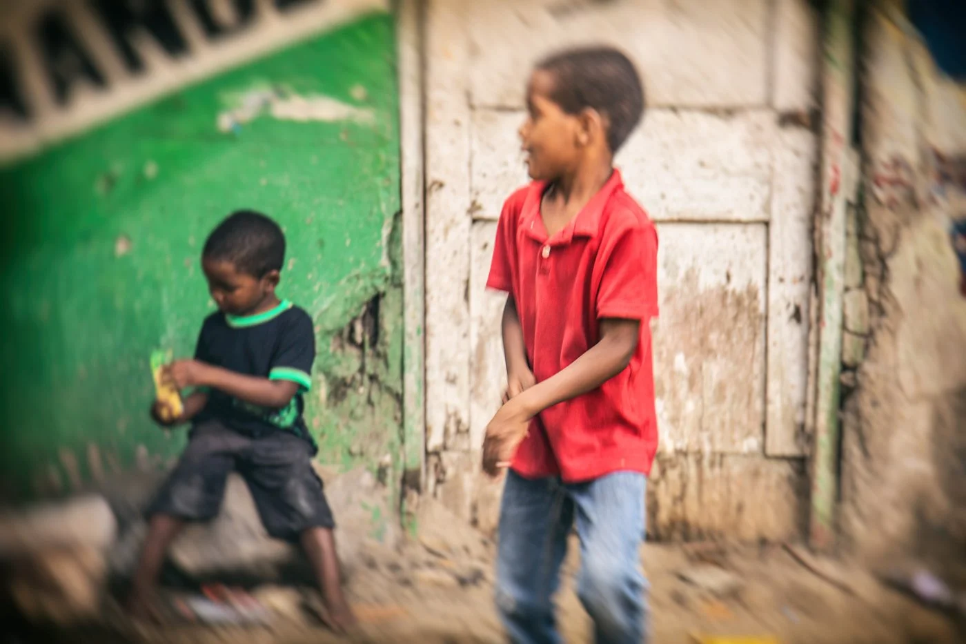 Two boys playing outside near a green wall and a wooden door in a rustic setting, one holding an object.