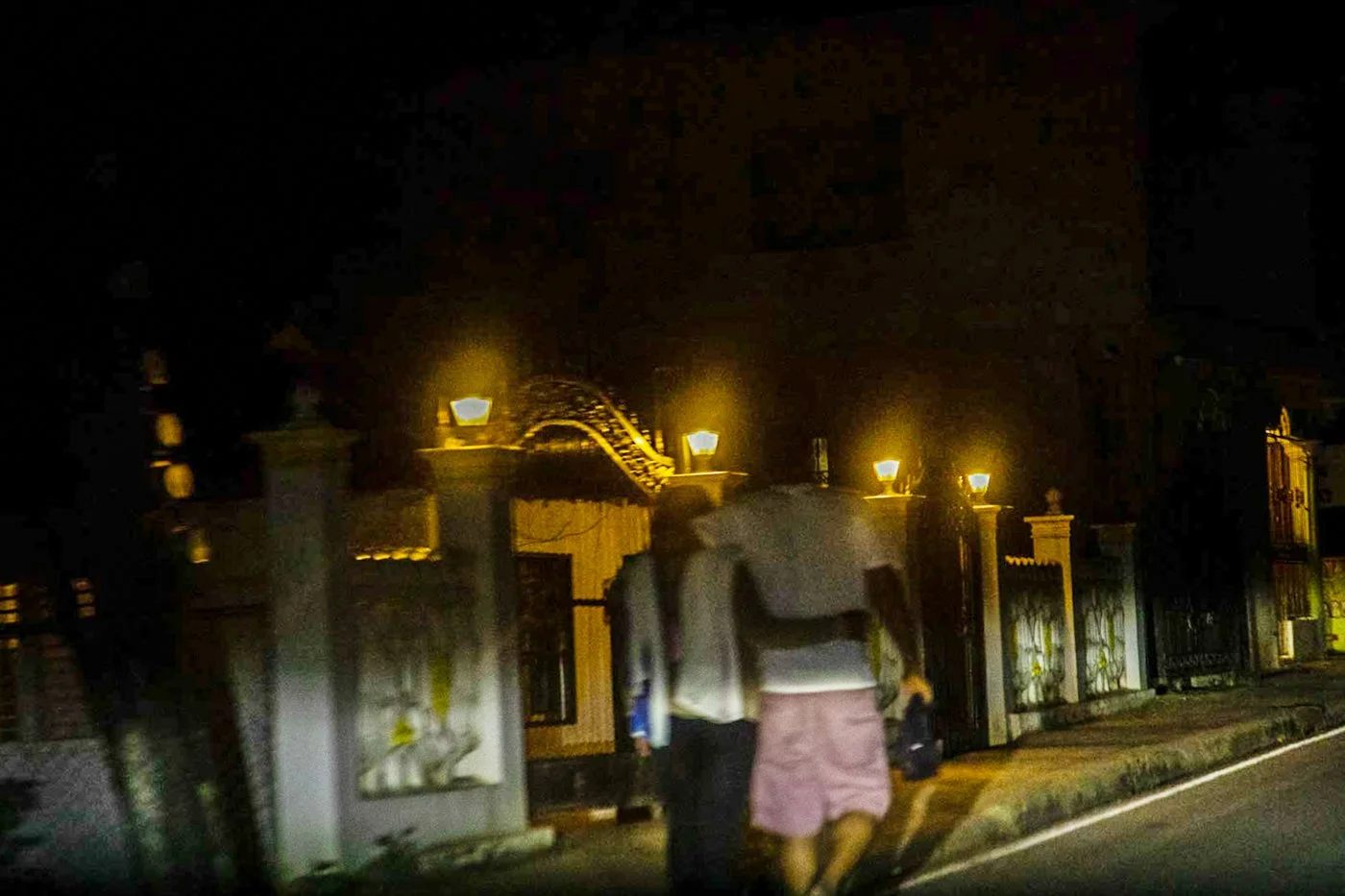 Nighttime scene of two people walking along a street near a house with decorative iron gate and illuminated lamps.