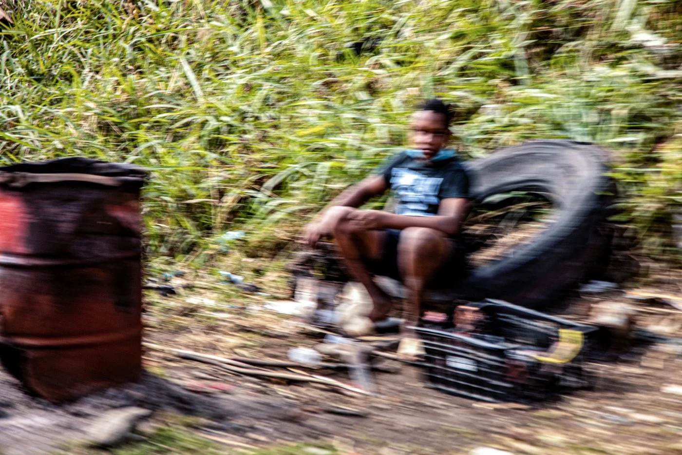 Blurred image of a person sitting next to a large tire and barrels in a grassy outdoor area.