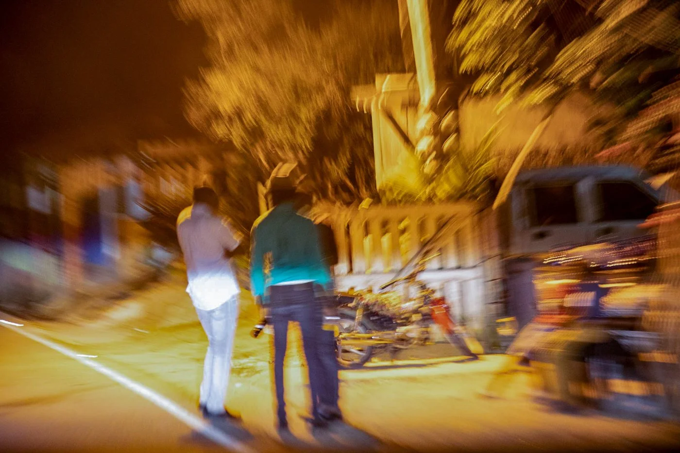 Blurry nighttime scene of two people standing on the street near a damaged vehicle and debris, with a truck parked nearby and illuminated by streetlights.