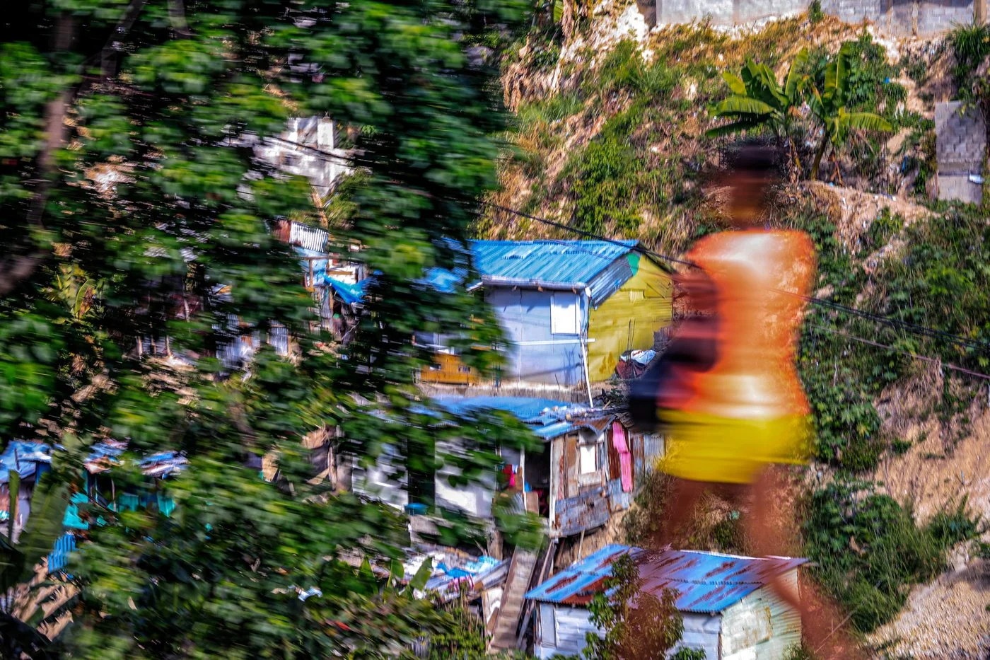 A blurry, colorful scene of a hillside with makeshift houses and a large, orange, dancing figure in the foreground.