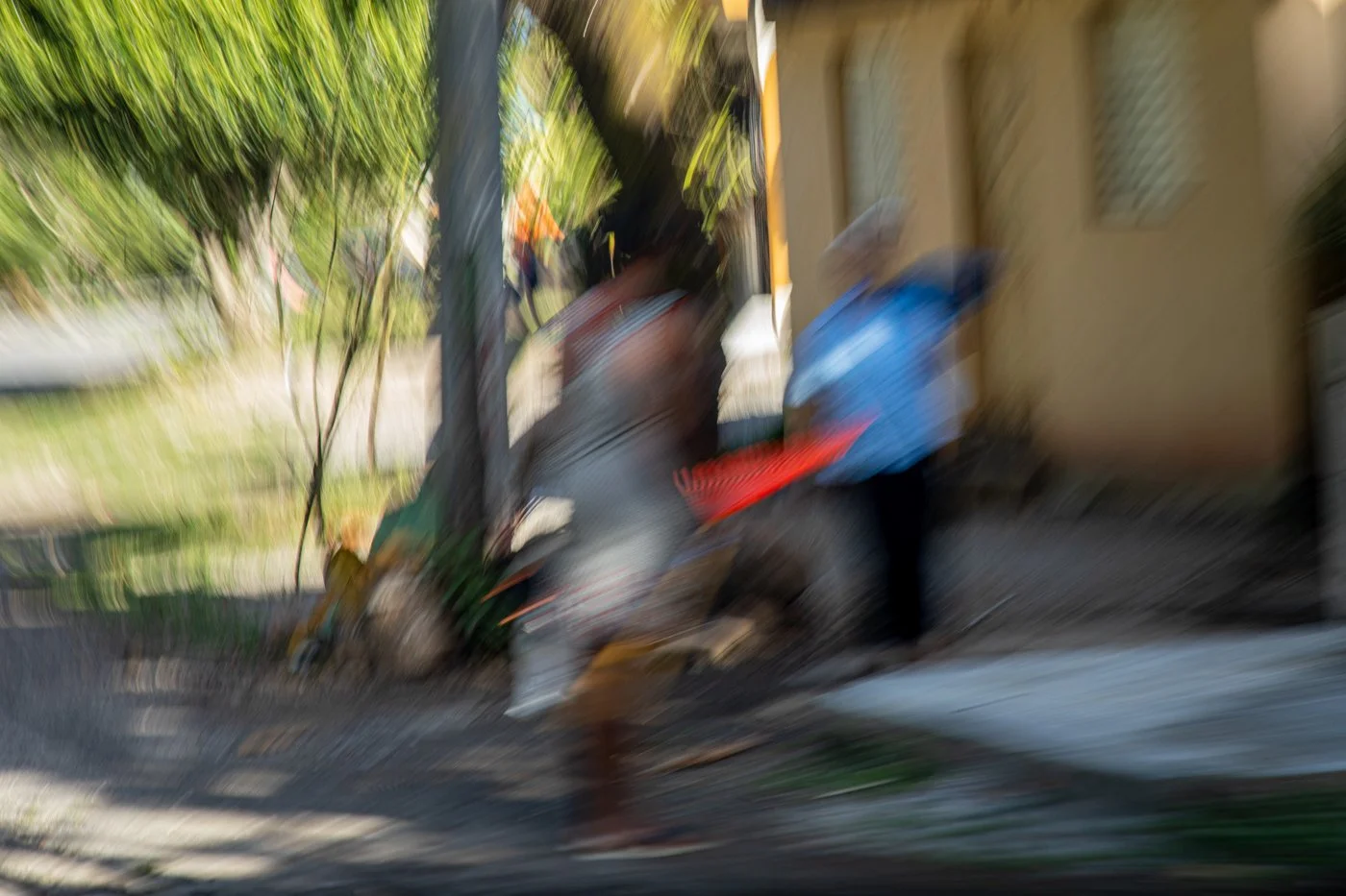 Blurry photo of two people walking along a sidewalk near a building with a yellow wall and windows, with trees and a fence in the background.