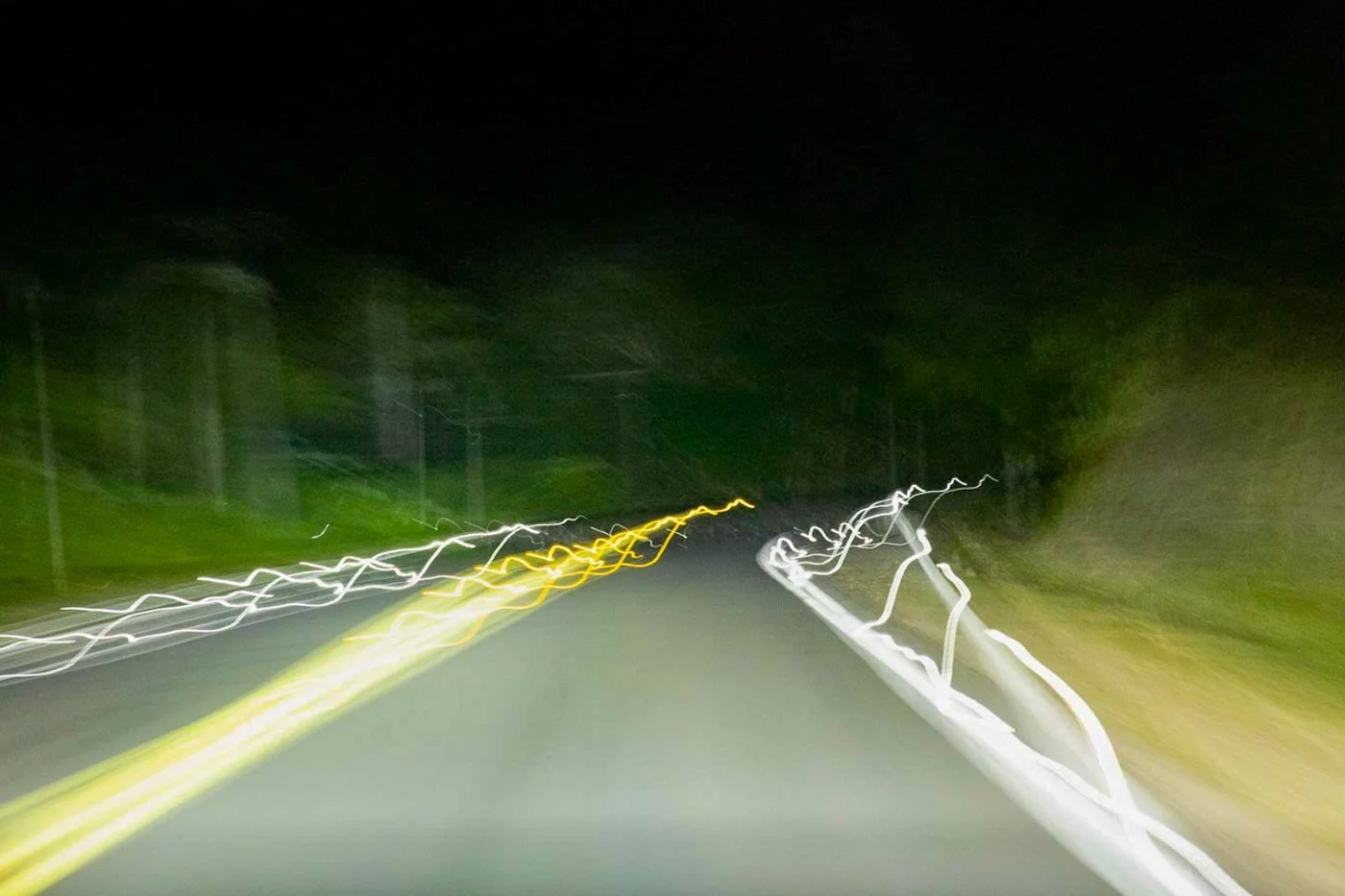 Nighttime road with blurred white and yellow light streaks due to camera motion, trees and road shoulder visible on sides.