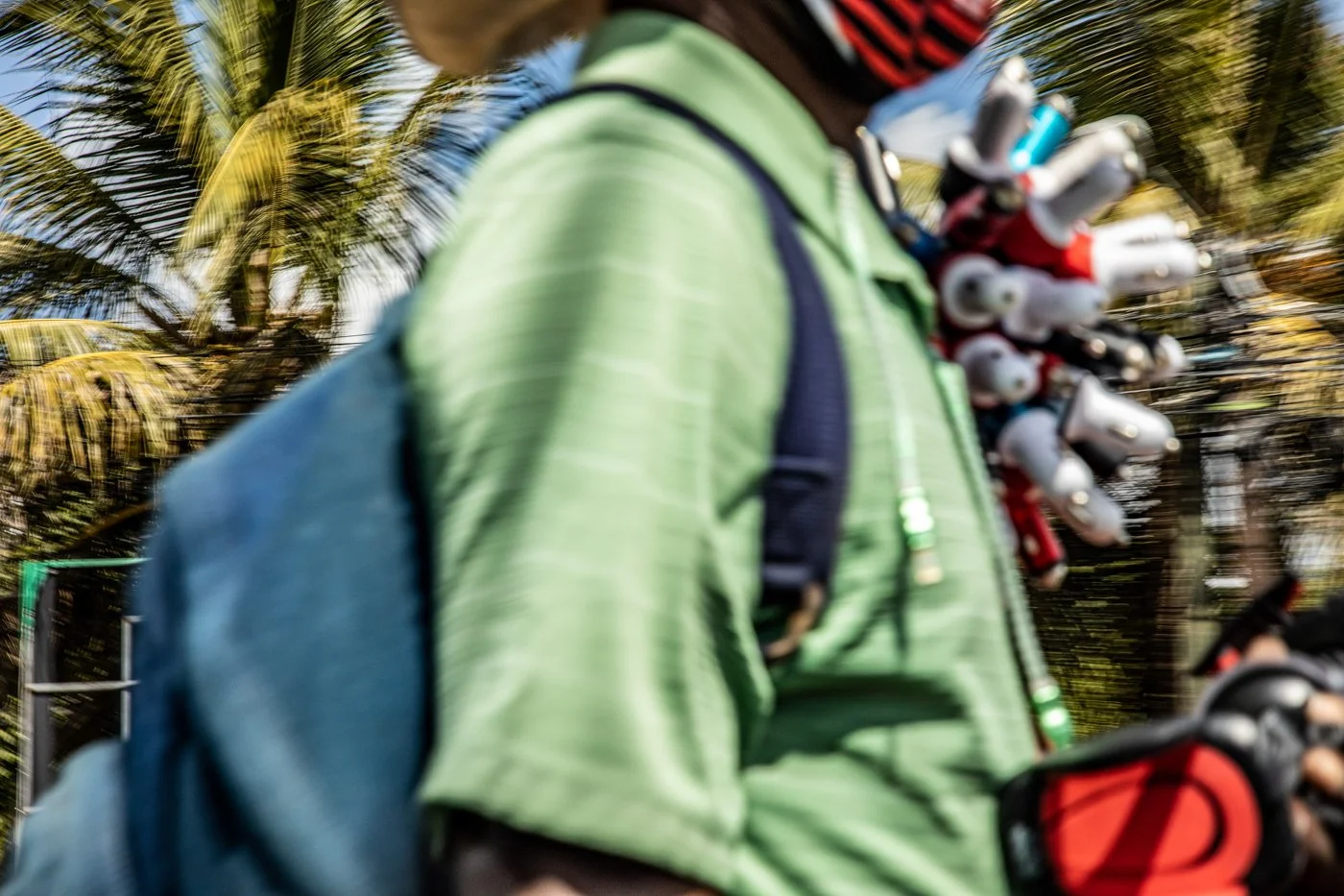 A person wearing a green jacket and carrying multiple tattoo machines around their neck, with palm trees and a sunny sky in the background.