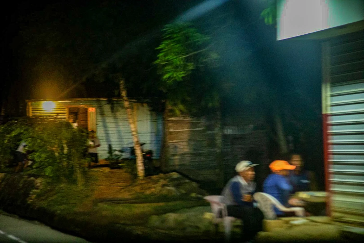 Nighttime street scene with two men sitting on plastic chairs near a building with a metal shutter, blurry from motion, and a small house or shed illuminated by a dim light in the background.