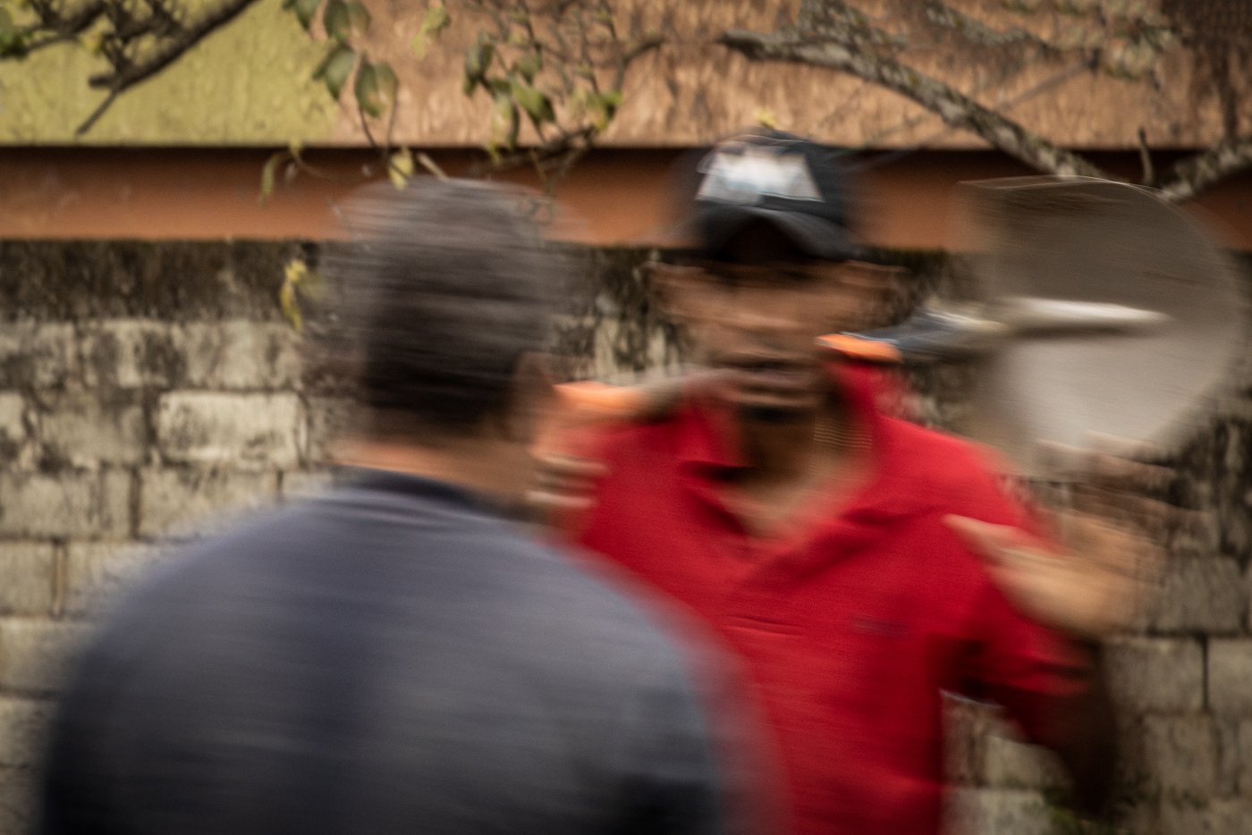 Two men engaged in a conversation in front of a brick wall, one wearing a red shirt with a cap, and the other with dark hair and a gray shirt, blurred due to motion.