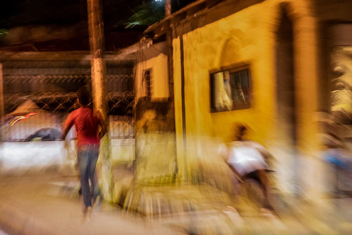 Two people on an urban street at night near a yellow building and a utility pole, with blurry motion and low lighting.