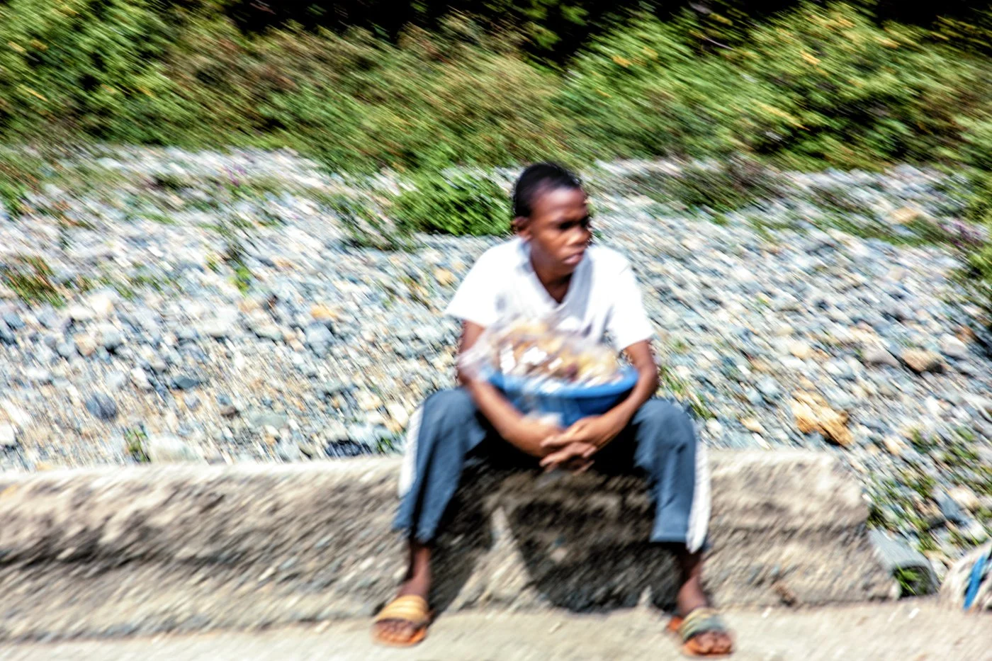 A person sitting on a concrete bench on a rocky and grassy area, holding a blue basket. The person is wearing a white shirt and gray pants with sandals. The background is blurred, creating a motion effect.
