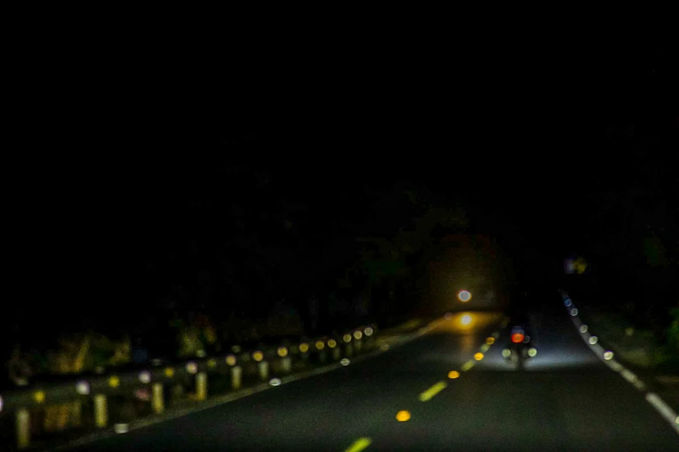 Nighttime road scene with two motorcyclists riding away from the camera, illuminated by their headlights, on a dark highway with reflective road markers.