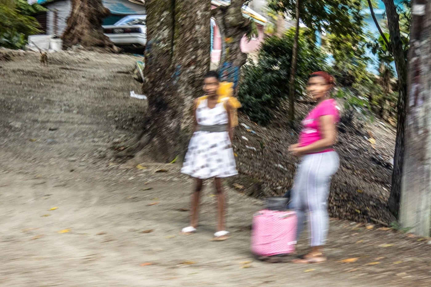 Two women standing on a dirt path near a large tree, carrying luggage and wearing casual clothes, in a rural area.