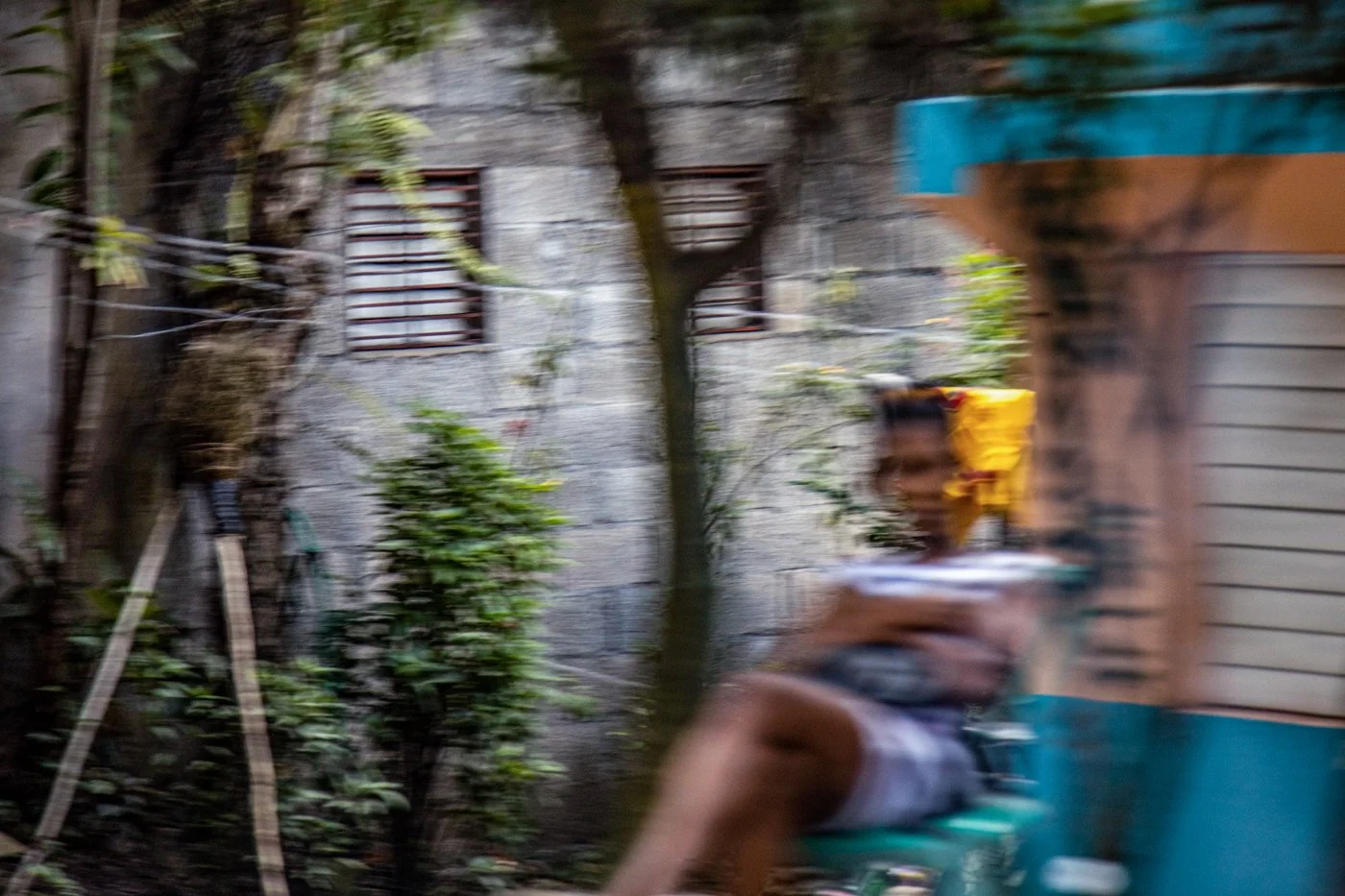 A blurry image of a person riding a motorcycle or scooter past a building with barred windows and plants.