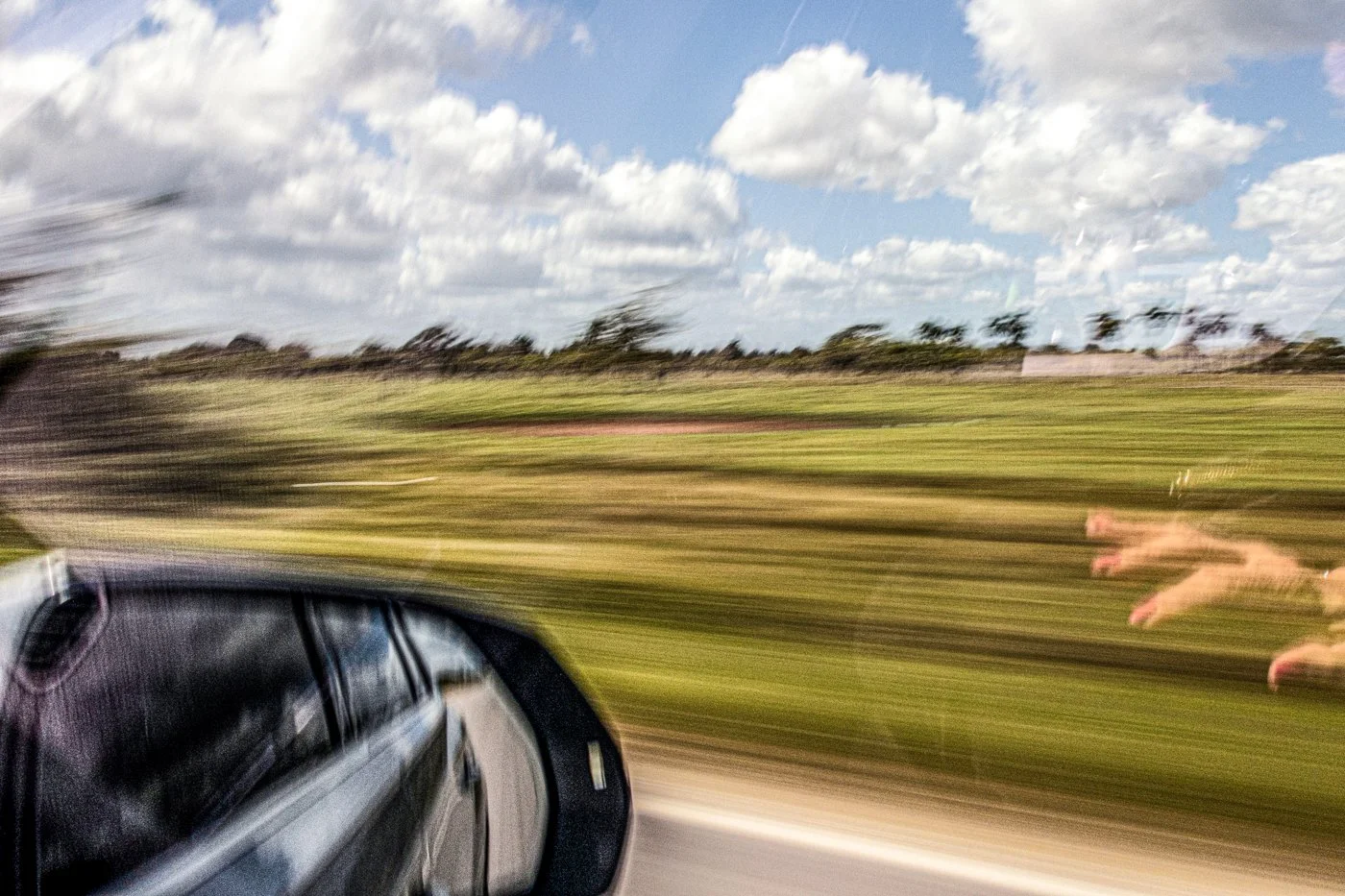 A blurred photo of a moving car's side mirror capturing a grassy landscape with trees and a partly cloudy sky, with a dog running alongside the moving vehicle.