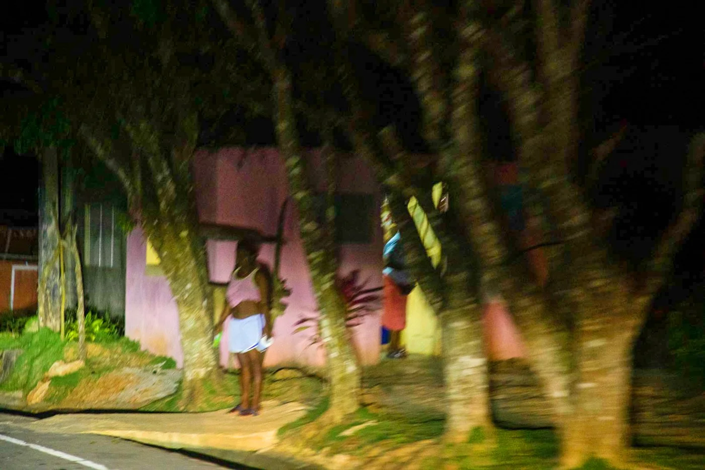 Nighttime street scene with several trees and a pink building in the background. A woman with dark skin, wearing a white dress standing on the sidewalk, and a person with a backpack near a tree.