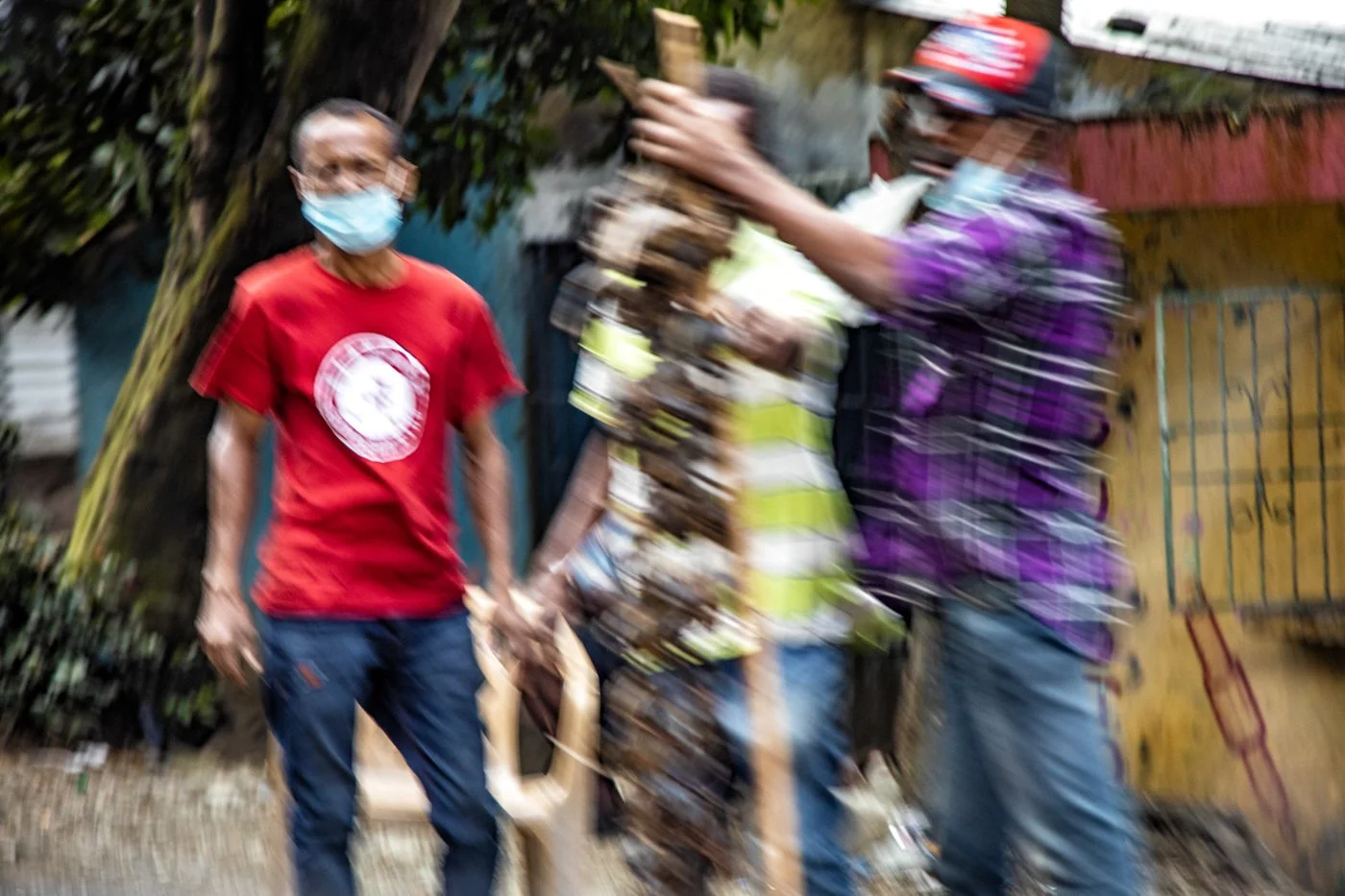A person in a red shirt and face mask stands outdoors with other individuals, one of whom is wearing a reflective vest, in a blurry scene.