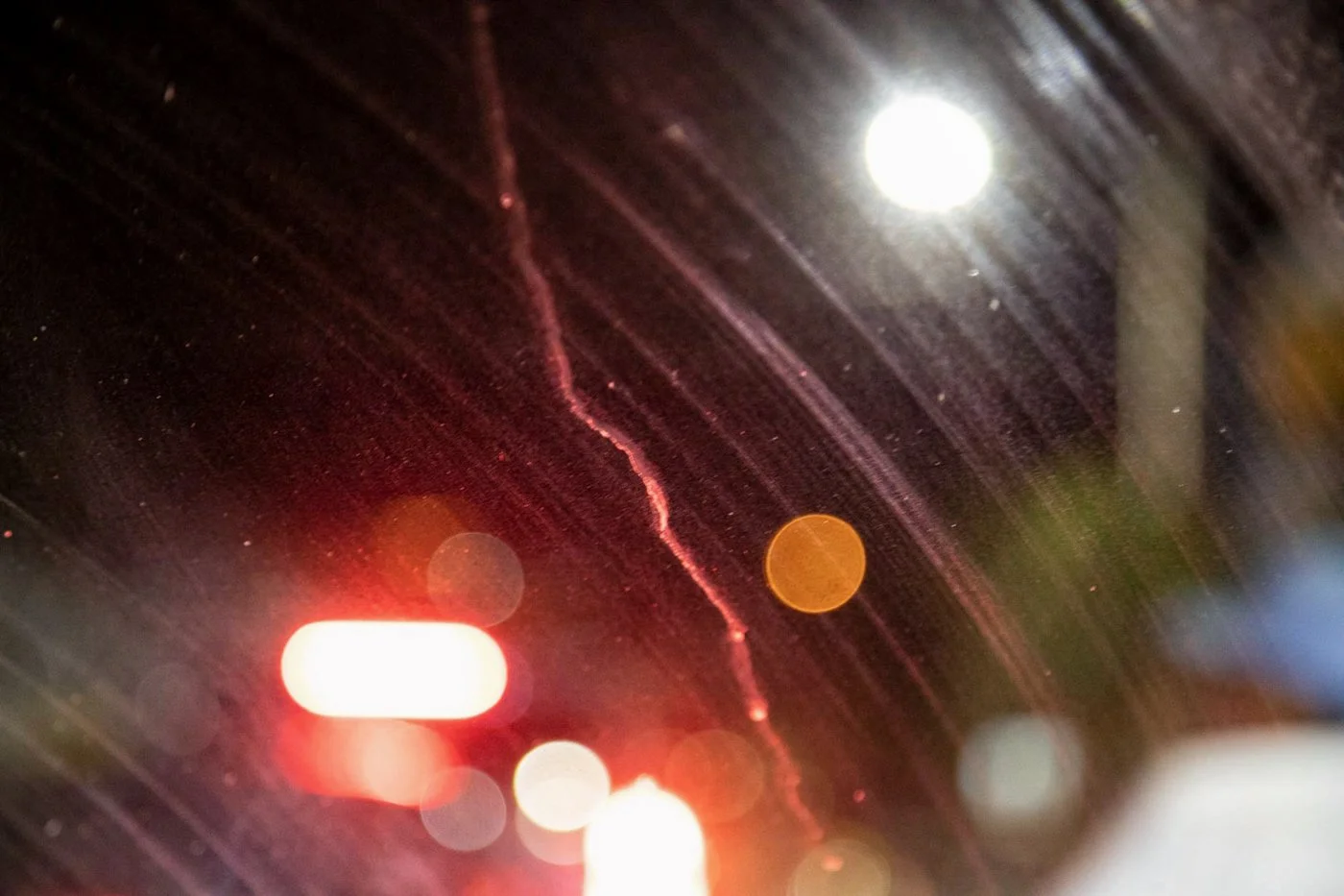 Close-up view of a windshield during a rainstorm at night, with visible streaks of rain, a bright streetlight, and blurred car lights creating a bokeh effect.